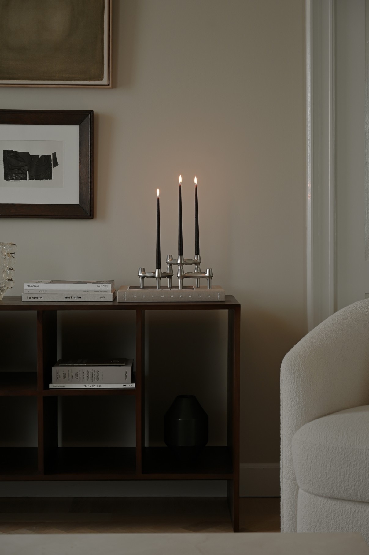 A lit three-candle candelabra on a wooden shelf next to an armchair, with framed art on the wall.
