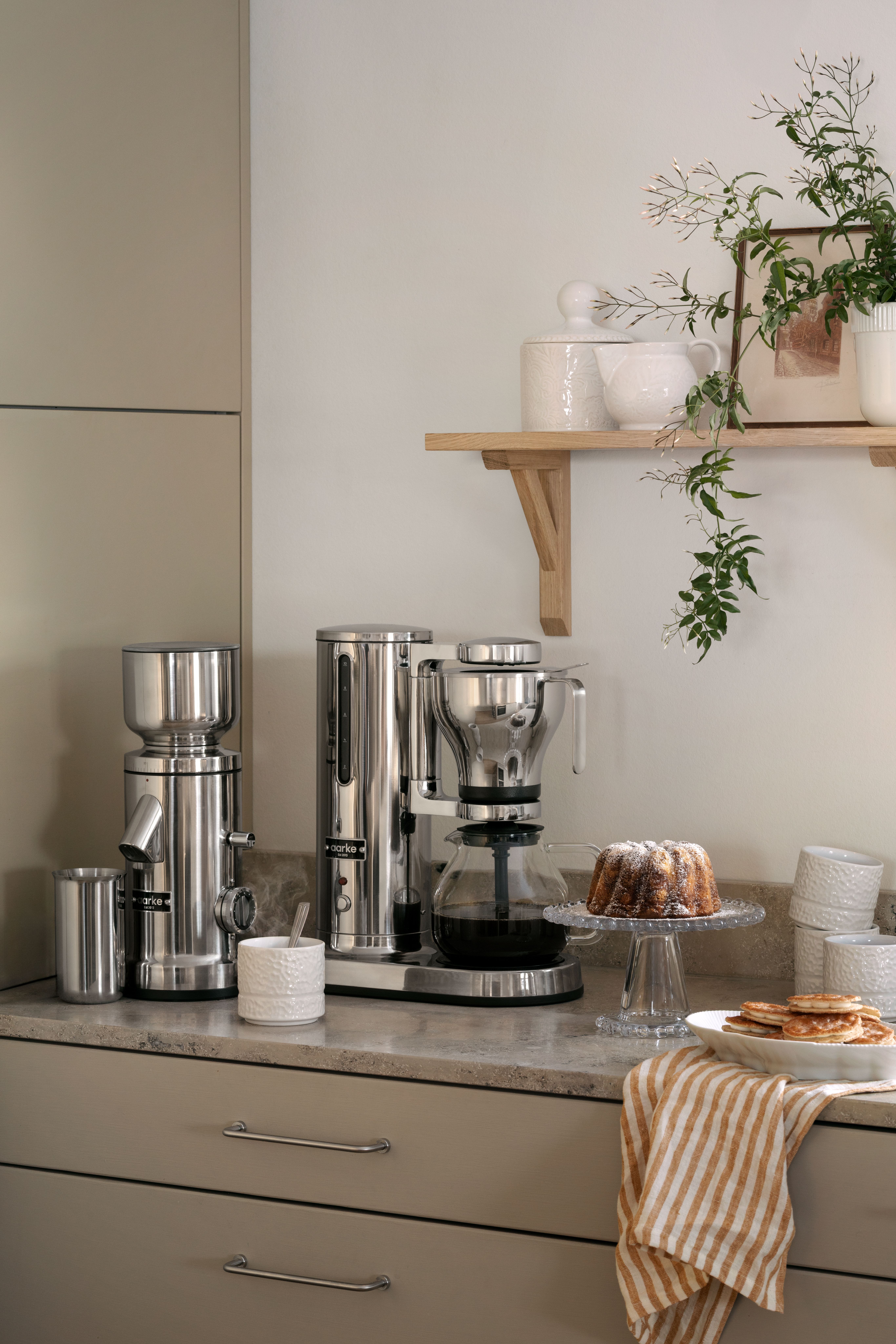 Kaffekvarn och kaffebryggare i rostfritt stål med en vit texturerad kanna på en köksbänk.