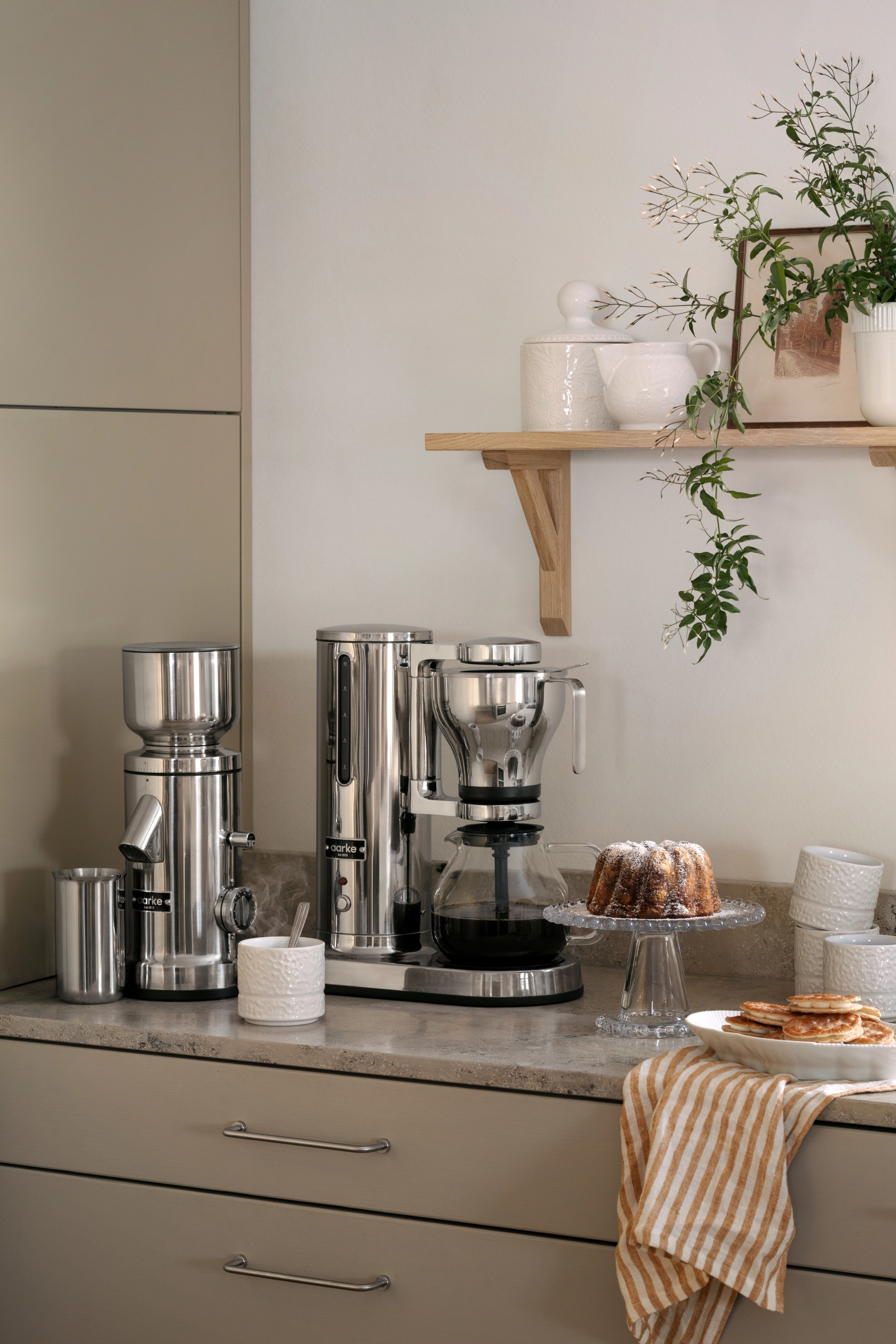 En modern köksbänk visar upp kaffemaskiner i rostfritt stål, en bundtkaka, pannkakor och en randig handduk, med en trähylla ovanför med växter och keramik.