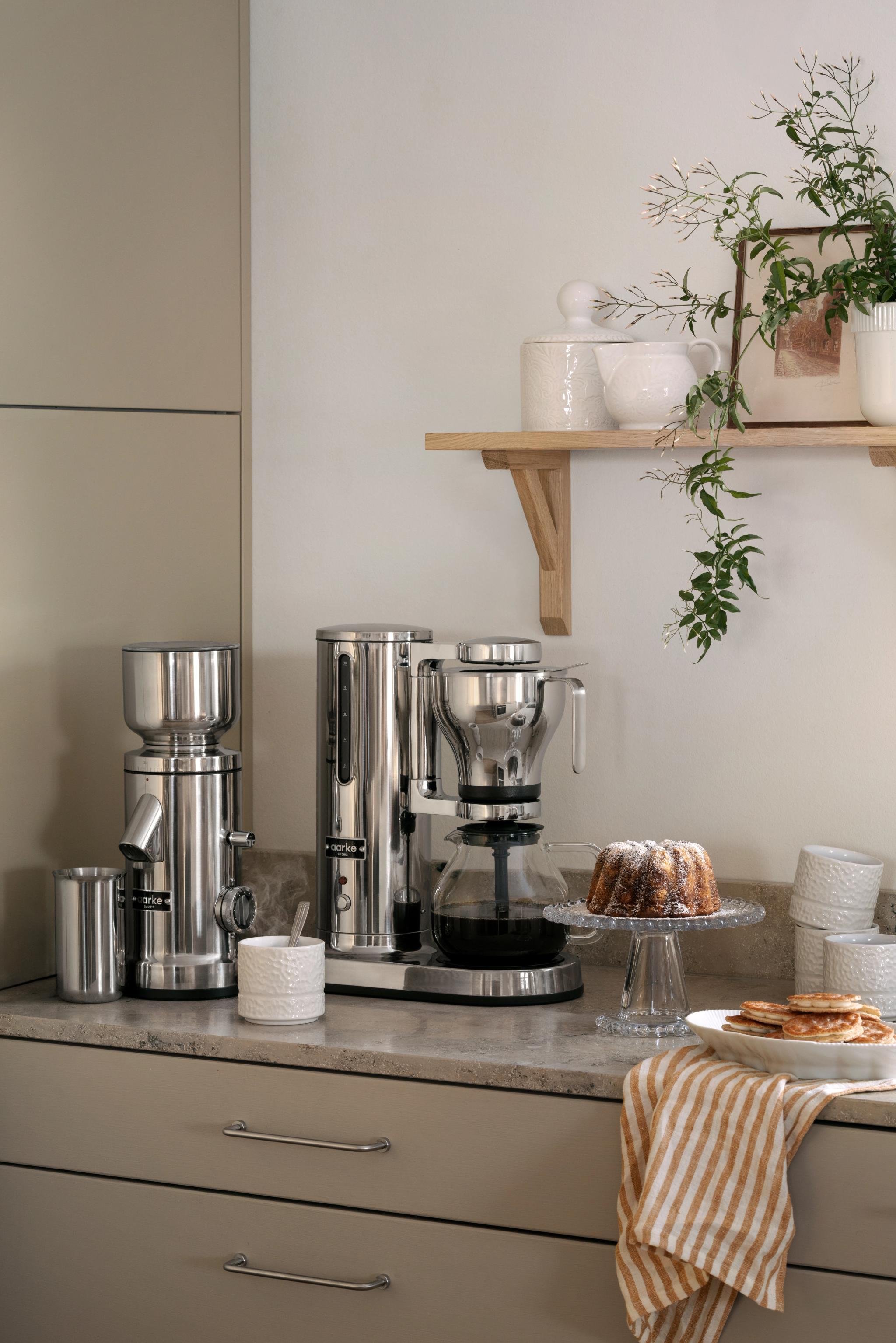 En modern köksbänk visar upp kaffemaskiner i rostfritt stål, en bundtkaka, pannkakor och en randig handduk, med en trähylla ovanför med växter och keramik.