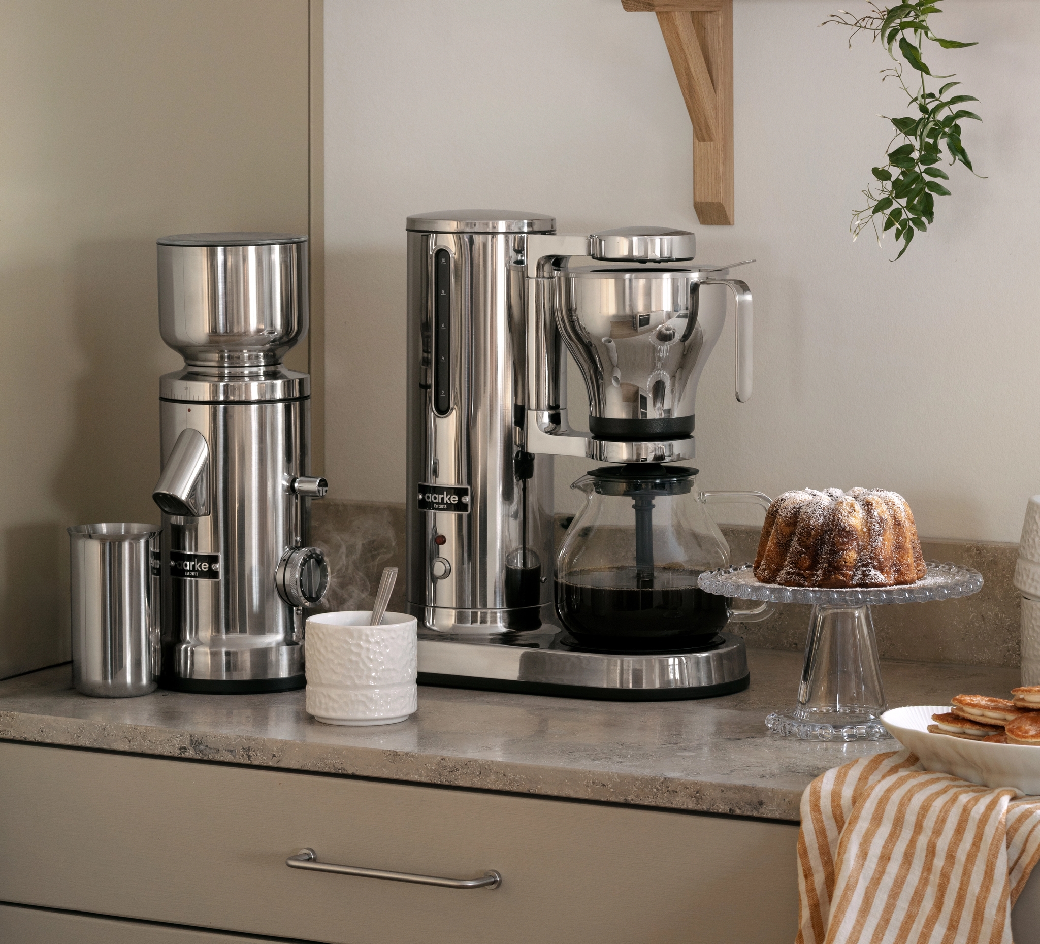 Kaffekvarn och kaffebryggare i rostfritt stål med en vit texturerad kanna på en köksbänk.