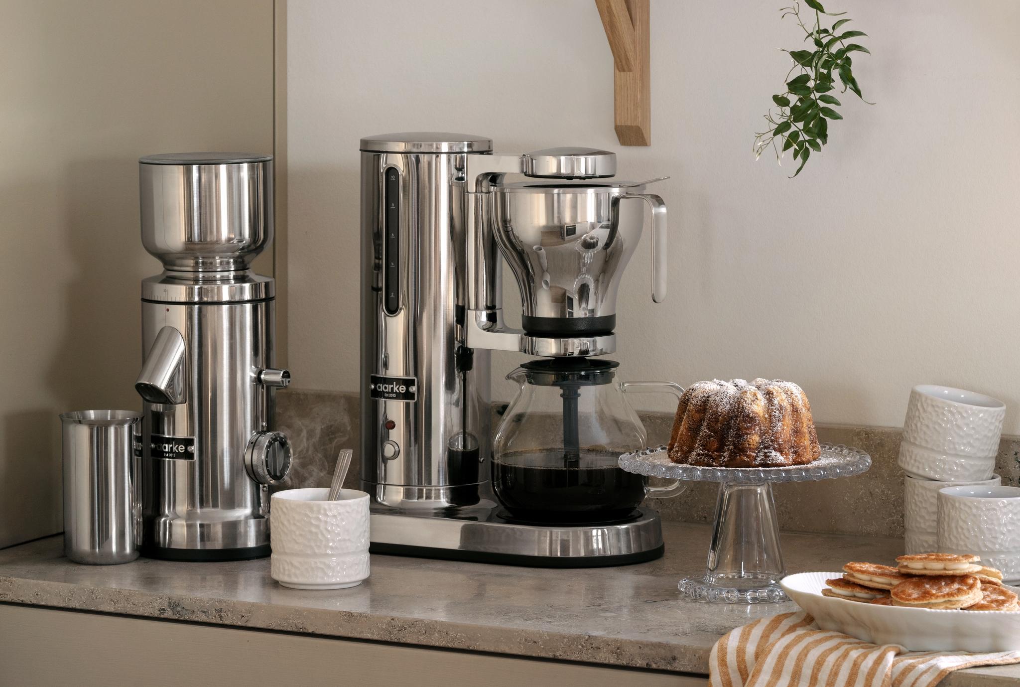 Kaffekvarn och kaffebryggare i rostfritt stål med en vit texturerad kanna på en köksbänk.