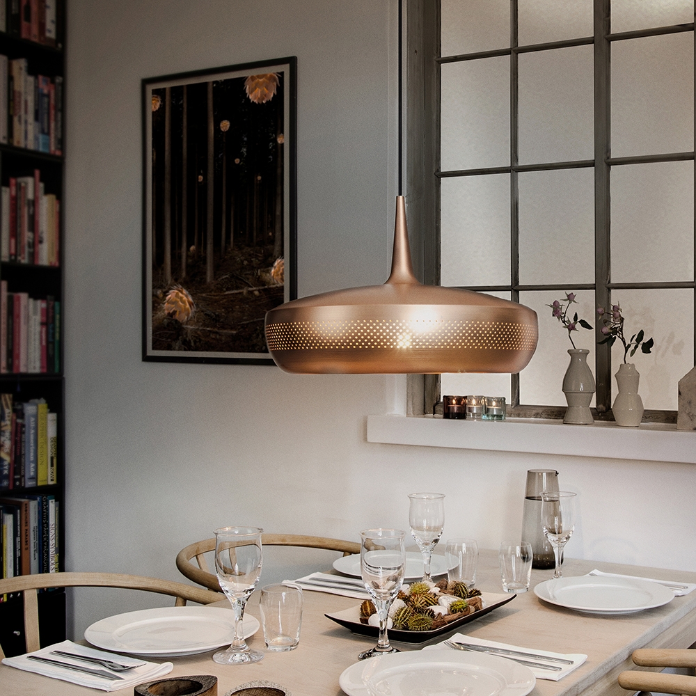 Matplats med ljus träbord, stilren dukning och modern kopparfärgad taklampa, omgiven av bokhylla, fönster med spröjs och dekorativa vaser i fönsternisch