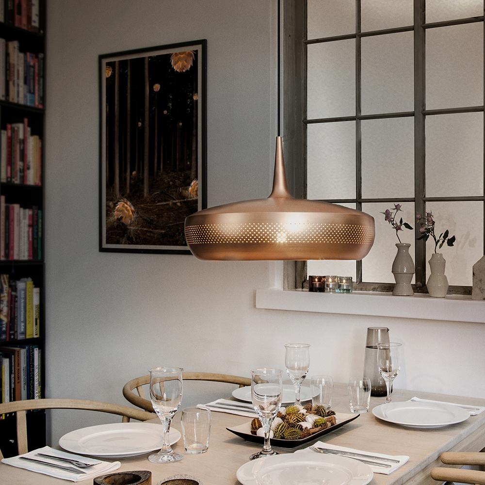 Matplats med ljus träbord, stilren dukning och modern kopparfärgad taklampa, omgiven av bokhylla, fönster med spröjs och dekorativa vaser i fönsternisch