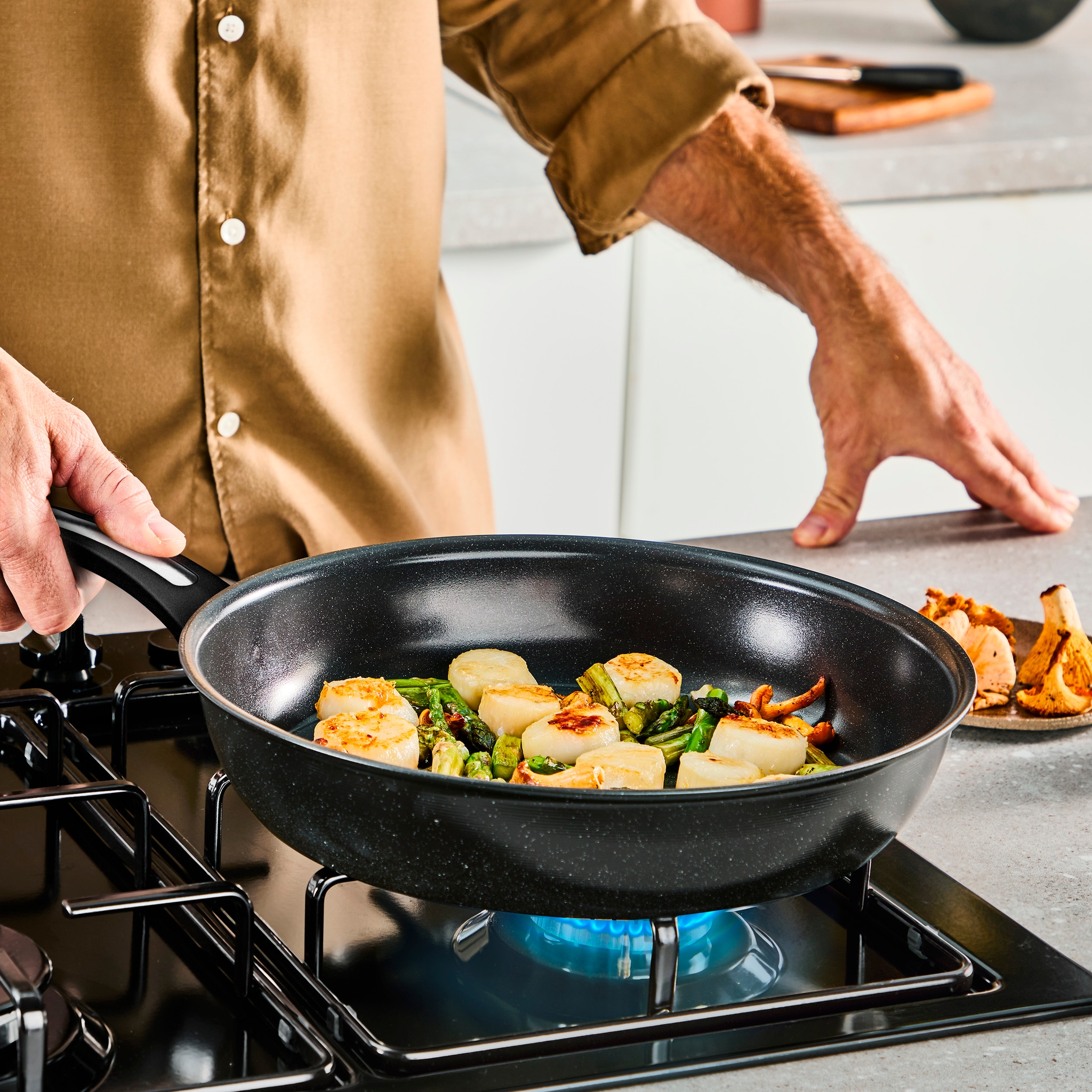 En person som steker kamskjell og asparges i en stekepanne på en gasskomfyr.