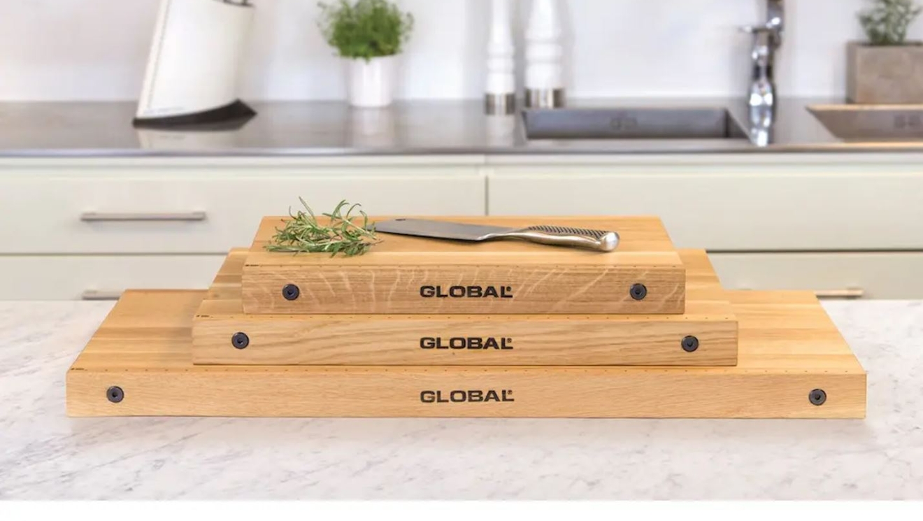 Global skärbräda i massivt trä i tre olika storlekar staplade på varandra, med Globalkniv och rosmarin ovanpå, placerade på marmorbänk i modernt kök