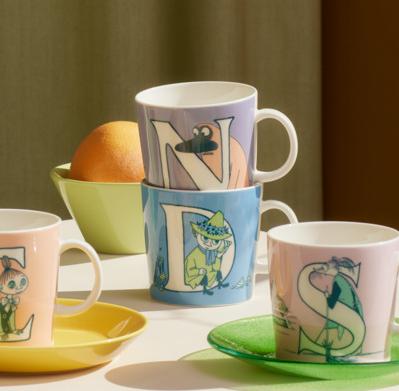 Färgglada muggar med bokstäver och illustrationer står arrangerade på ett bord i mjukt, varmt ljus.