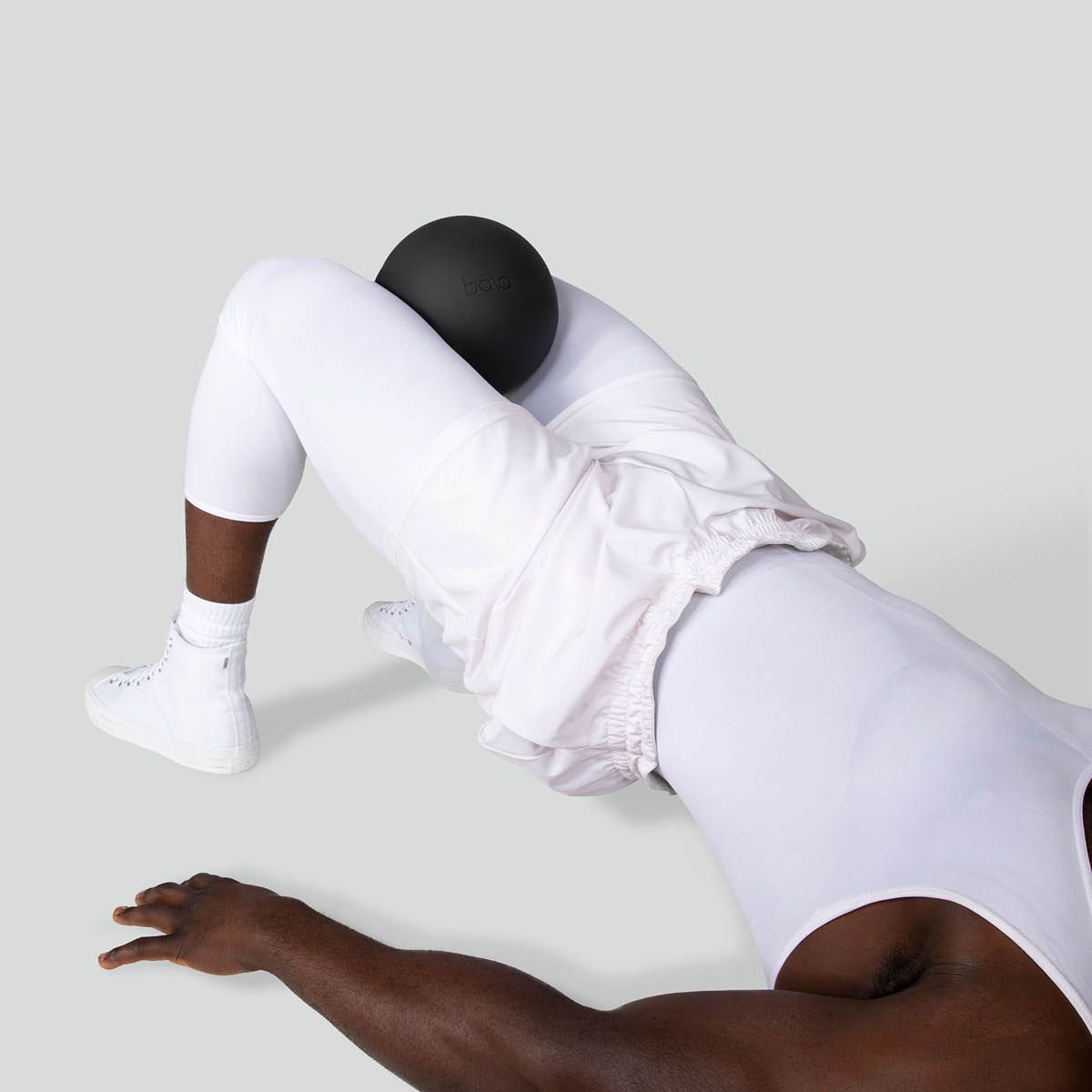 Valkoisiin urheiluvaatteisiin pukeutunut henkilö makaa selällään ja puristaa mustaa palloa koukistettujen polviensa välissä.