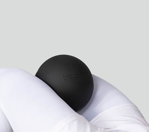 Valkoisiin urheiluvaatteisiin pukeutunut henkilö makaa selällään ja puristaa mustaa palloa koukistettujen polviensa välissä.