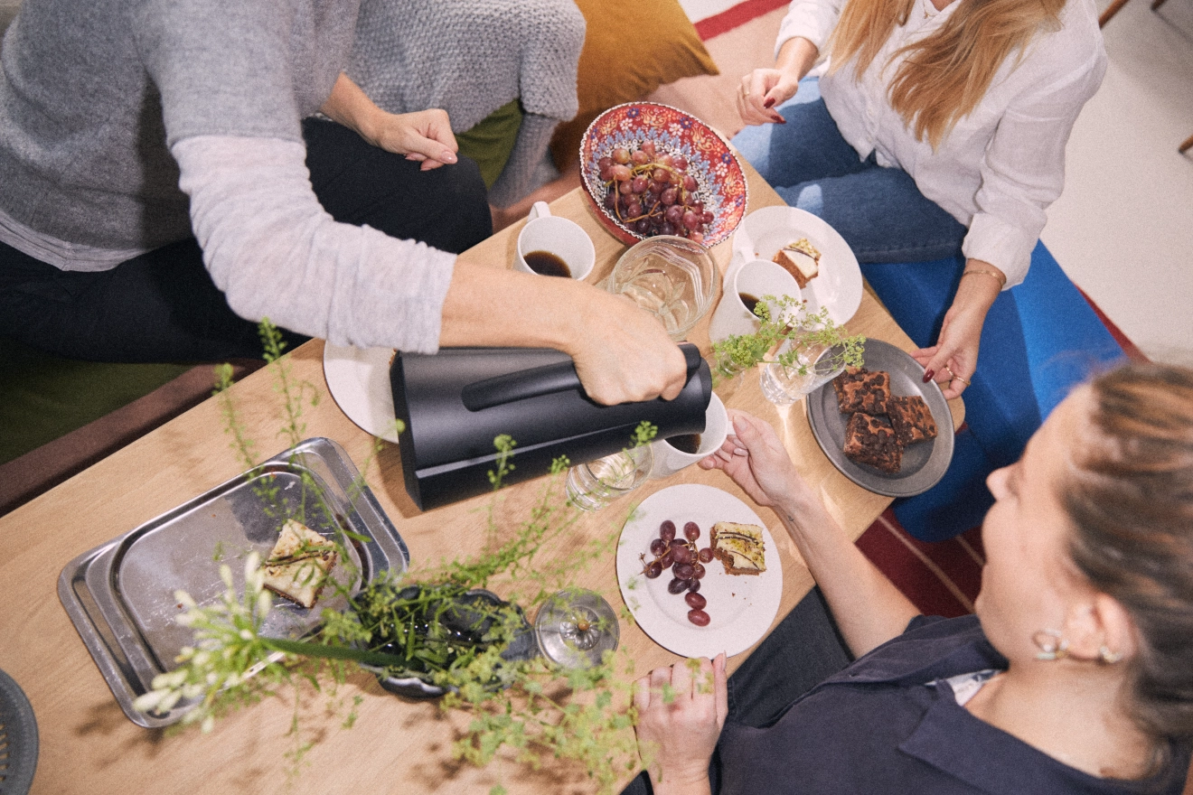 Ylhäältä kuvattu näkymä ihmisistä pöydän ympärillä, jossa on kahvia, viinirypäleitä, brownie-leivoksia ja jälkiruokia; yksi henkilö kaataa kahvia