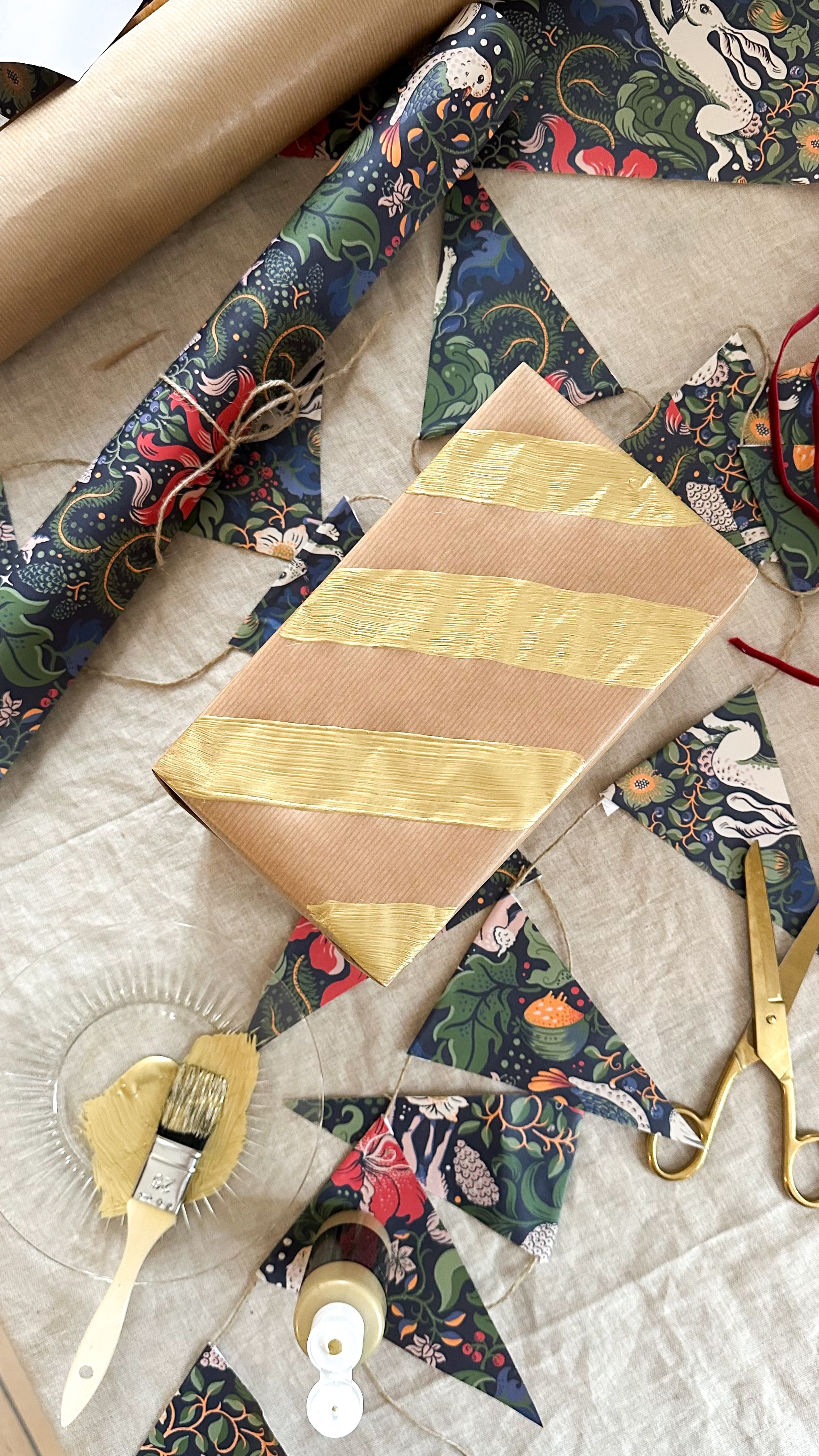 A brown paper gift with gold stripes, patterned wrapping paper, bunting, gold paint, and scissors arranged on a linen surface.