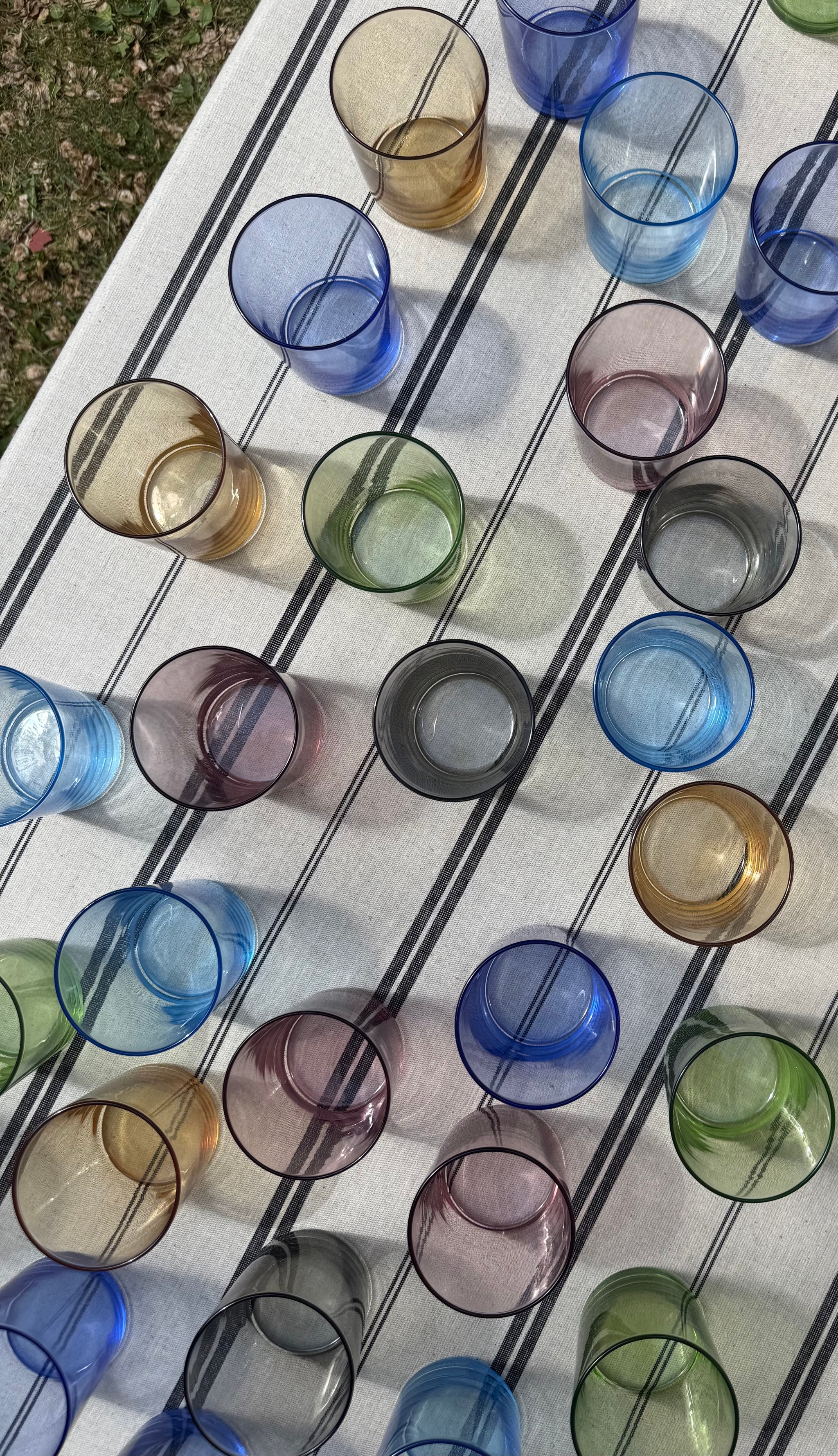 Färgglada dricksglas i olika nyanser står utspridda på en vit duk med svarta ränder utomhus.