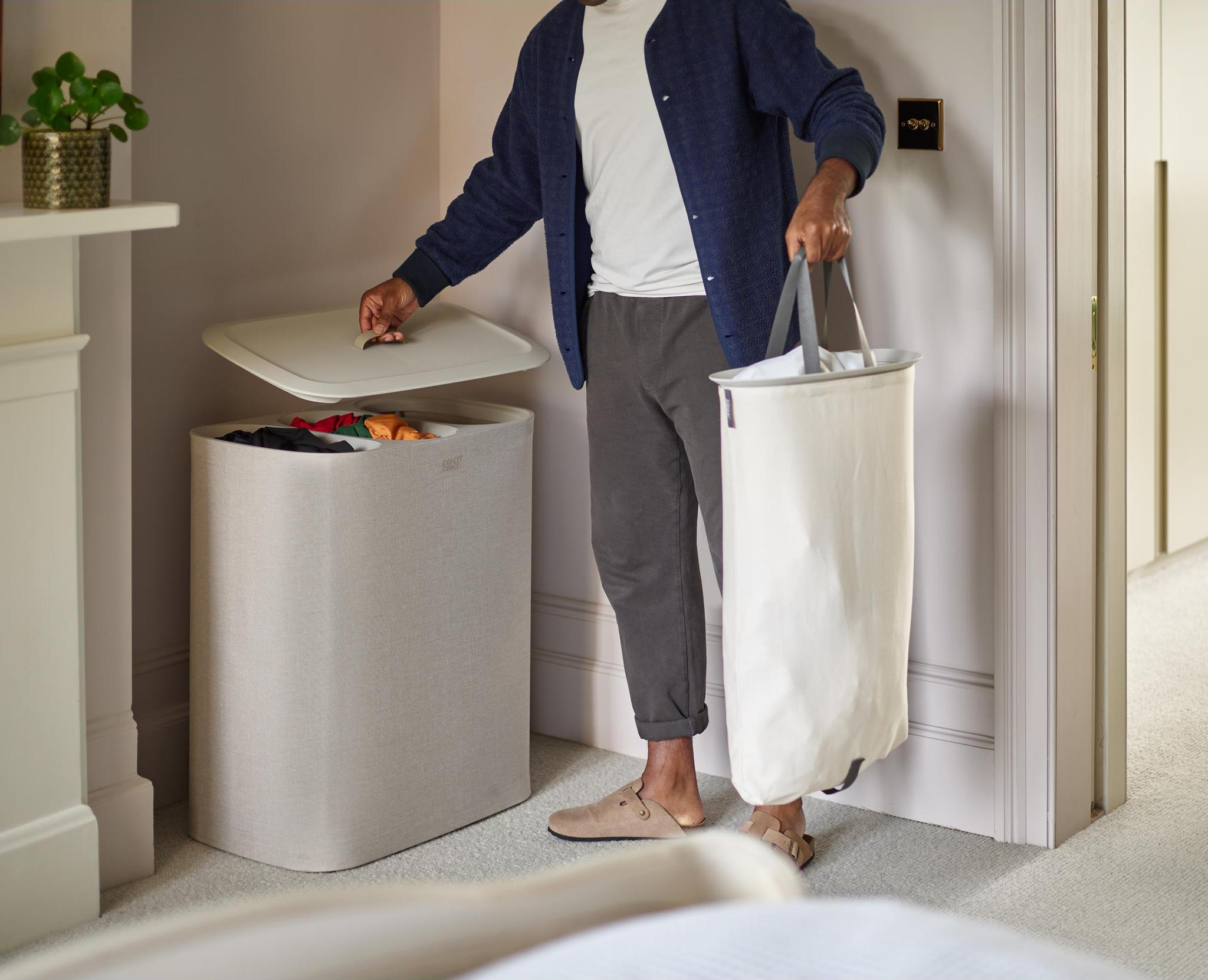 Person lägger tvätt i en stilren, beige tvättkorg med lock i ett ljust sovrum.