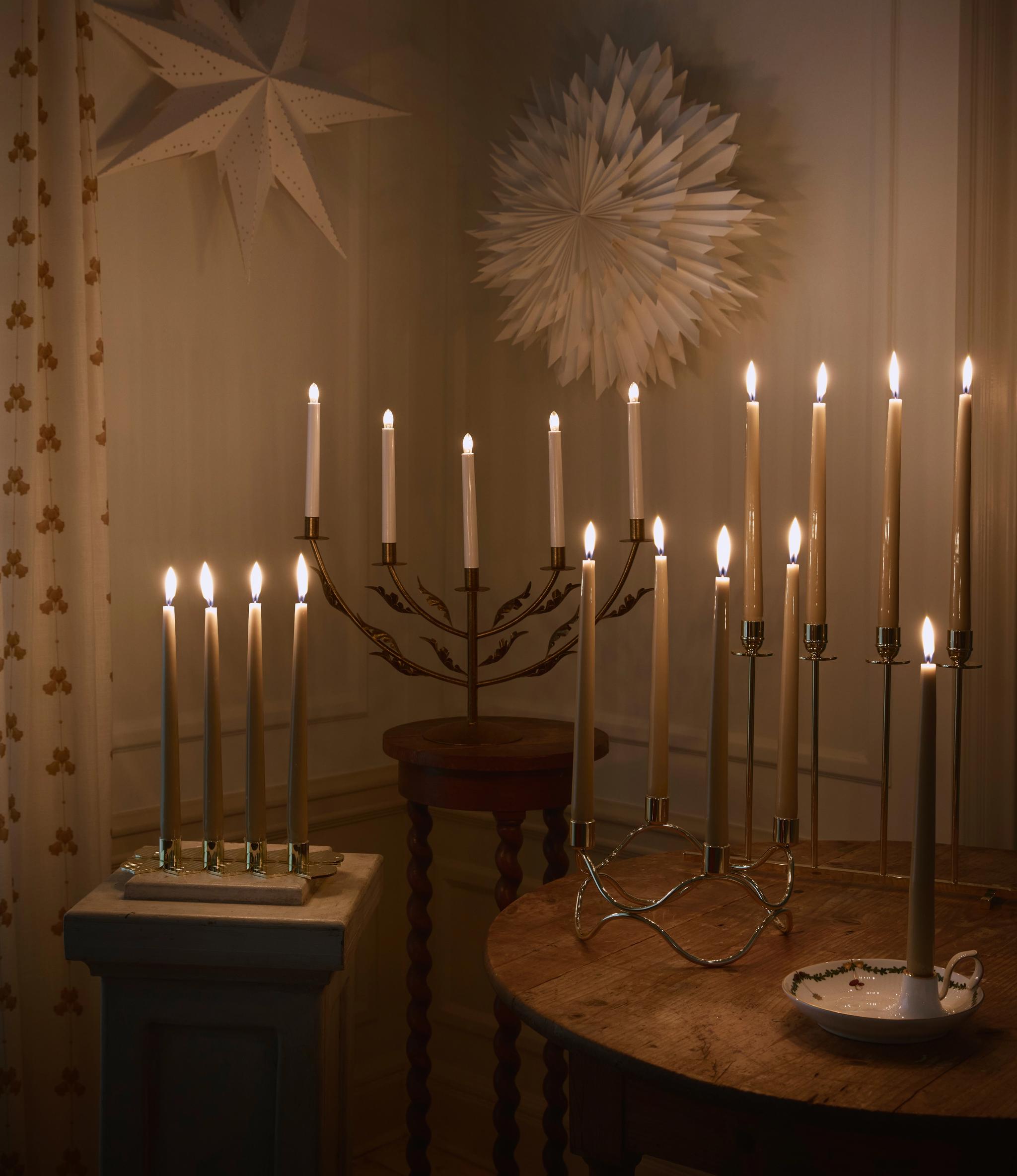 Lämmin huone, joka on täynnä palavia kynttilöitä erilaisissa kynttilänjaloissa, sekä kaksi paperitähteä seinällä ja puisia pöytiä.