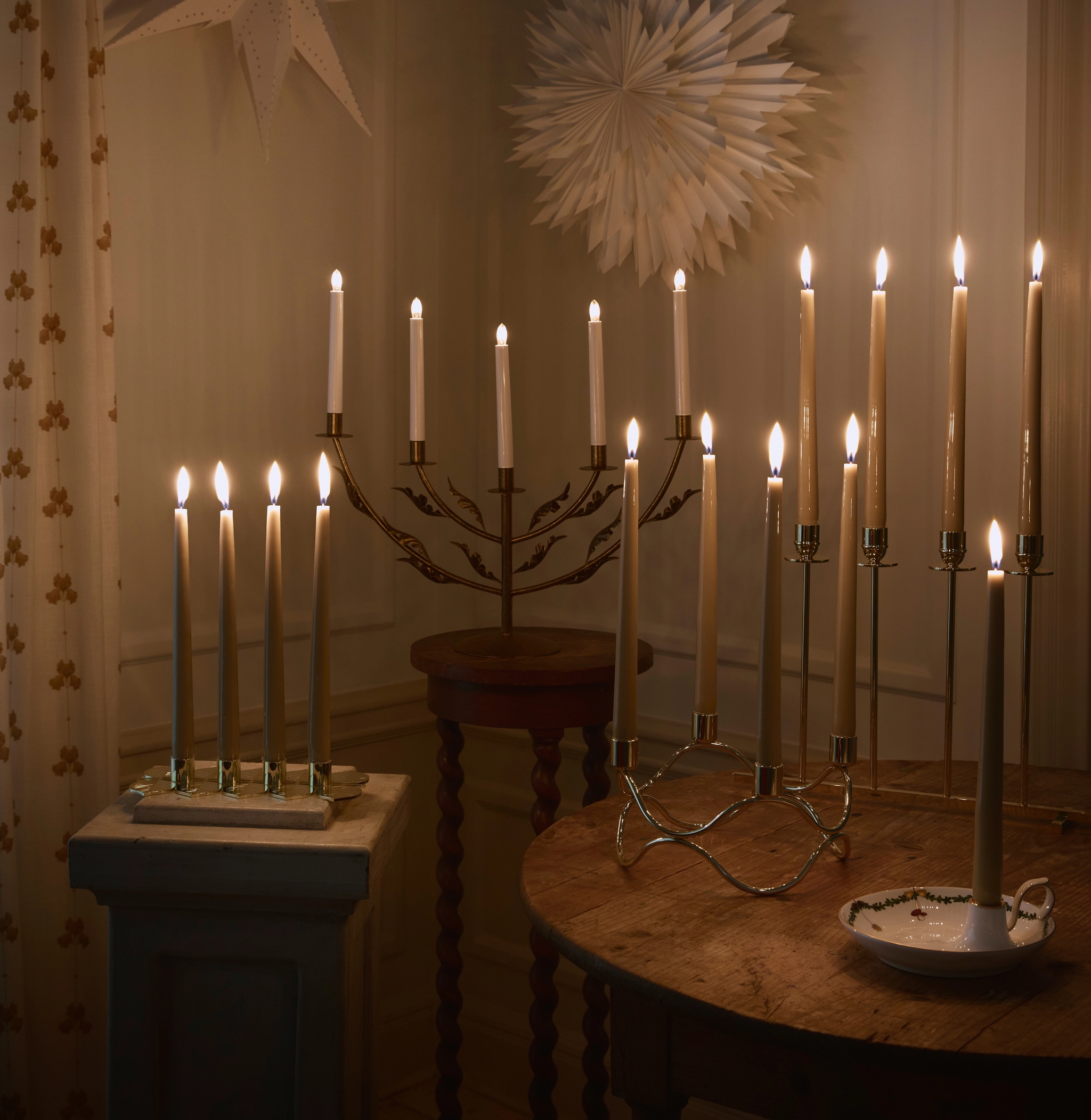 Lämmin huone, joka on täynnä palavia kynttilöitä erilaisissa kynttilänjaloissa, sekä kaksi paperitähteä seinällä ja puisia pöytiä.