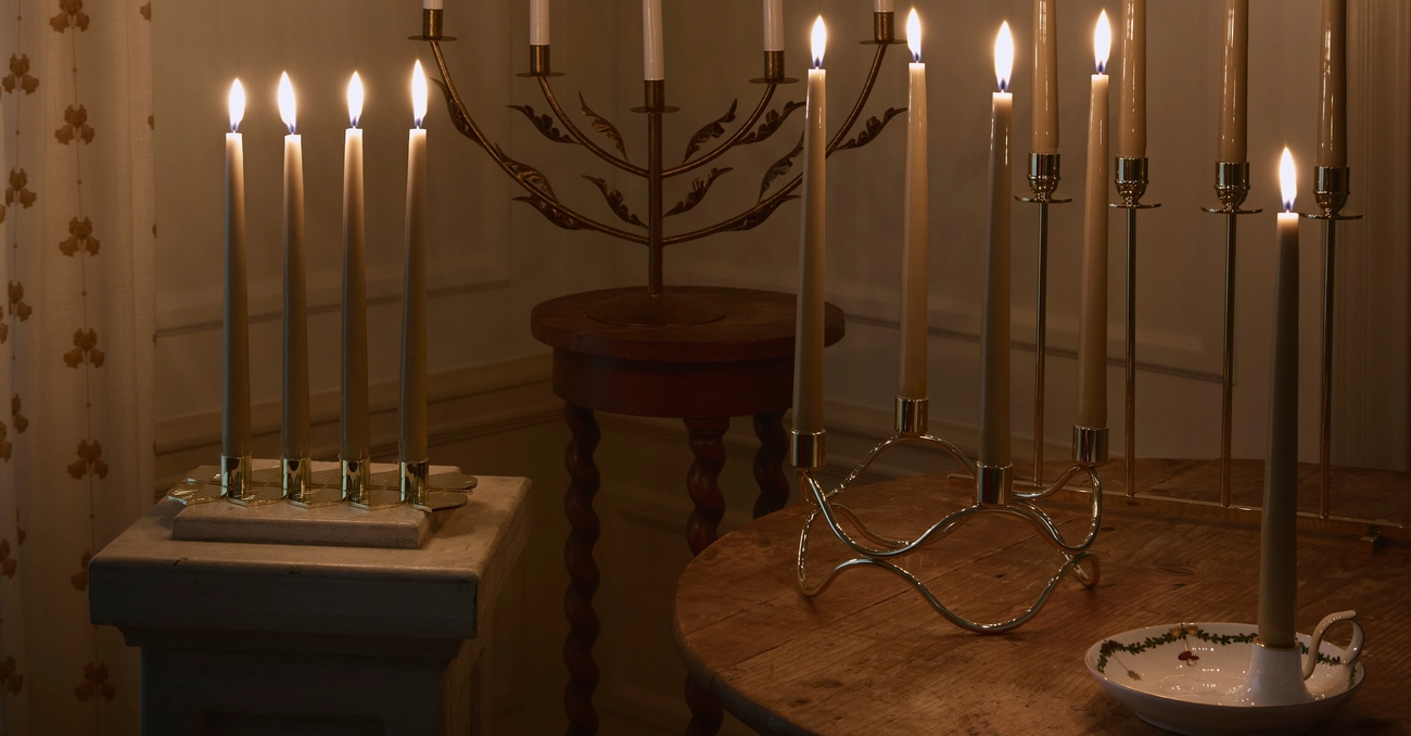 Lämminhenkinen huone, jonka täyttää lukuisten palavien kynttilöiden pehmeä hehku. Kynttilät on aseteltu eri tasoille ja pöydille, ja seinällä loistaa kaksi koristeellista paperitähteä.