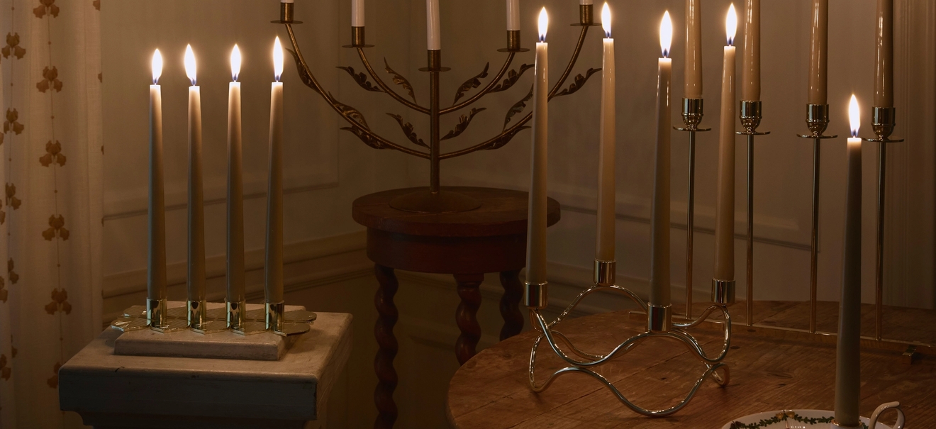 Lämminhenkinen huone, jonka täyttää lukuisten palavien kynttilöiden pehmeä hehku. Kynttilät on aseteltu eri tasoille ja pöydille, ja seinällä loistaa kaksi koristeellista paperitähteä.