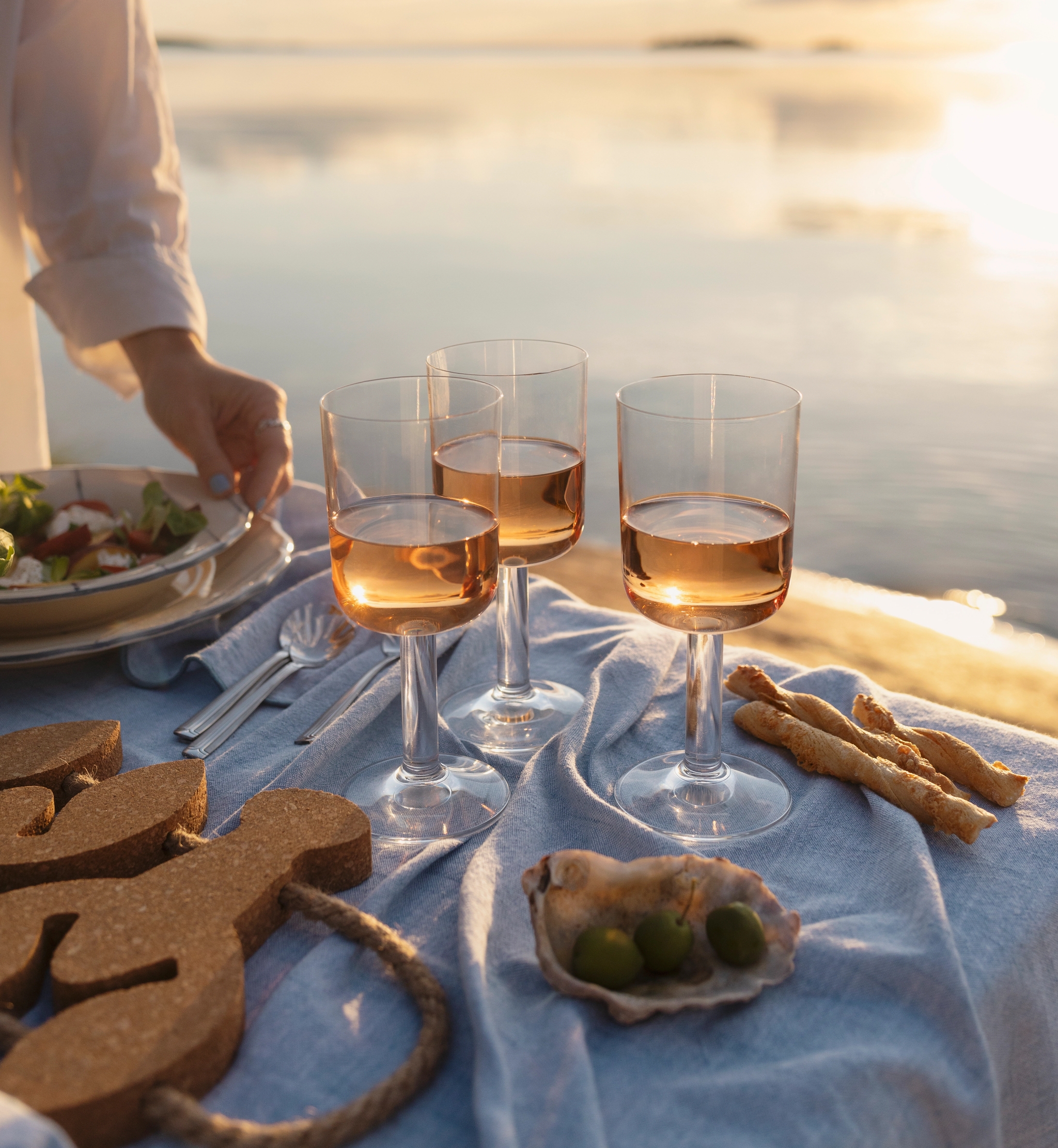 Somrig picknickdukning med rosévin, snacks och sallad vid en stilla sjö i solnedgången.