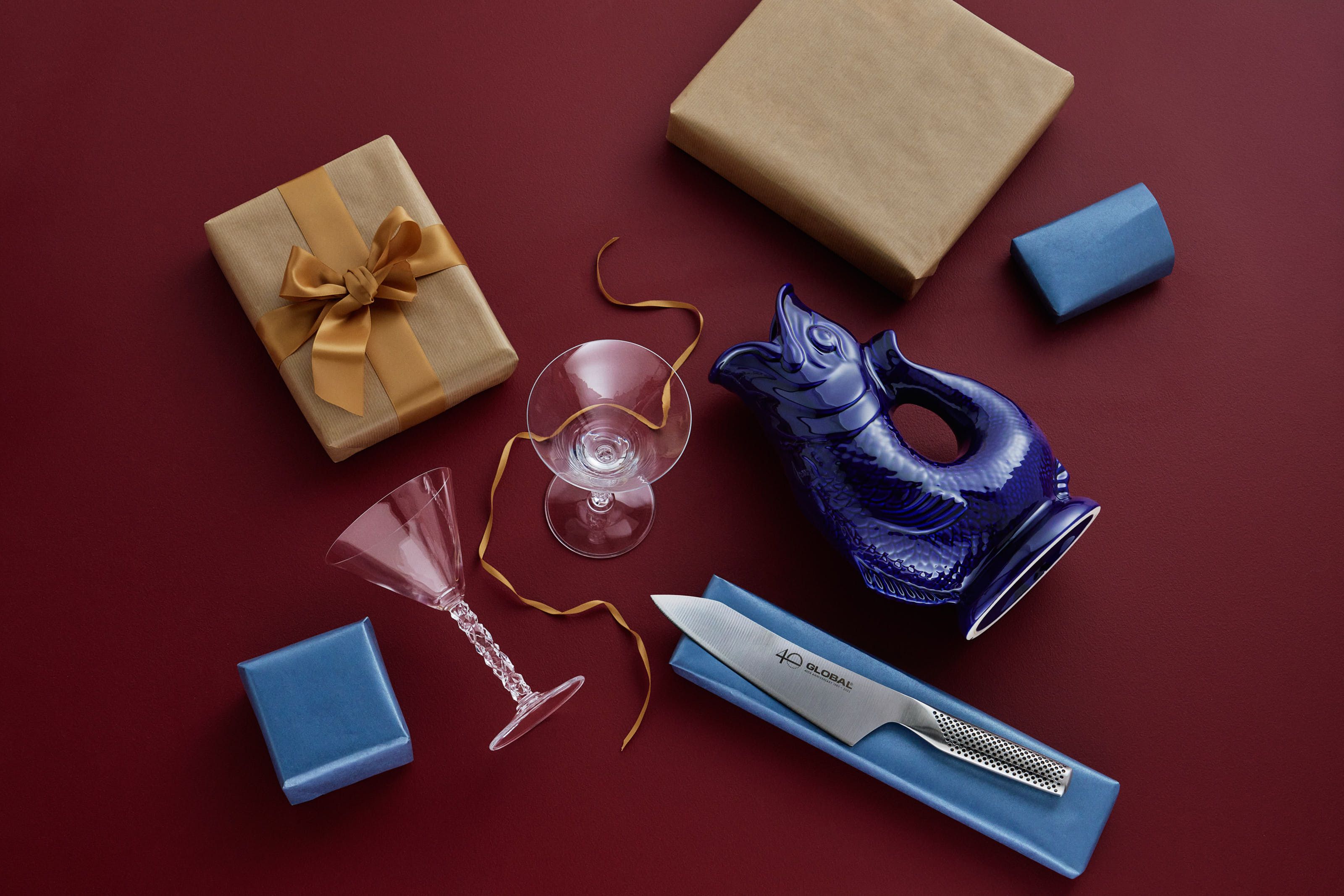 En bild ovanifrån av presenter och hushållsartiklar, inklusive inslagna lådor, en blå fiskkanna, en kockkniv och glas, mot en röd bakgrund.