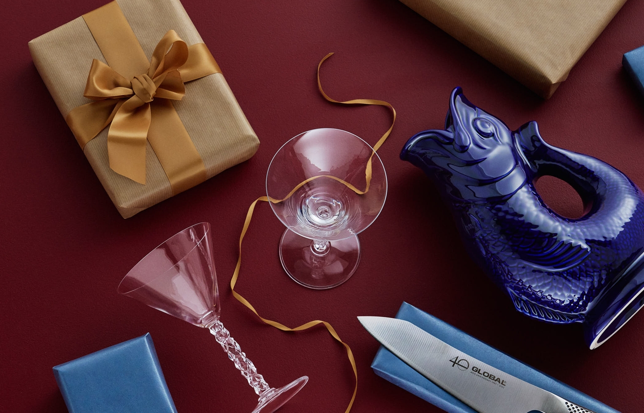 En bild ovanifrån av presenter och hushållsartiklar, inklusive inslagna lådor, en blå fiskkanna, en kockkniv och glas, mot en röd bakgrund.