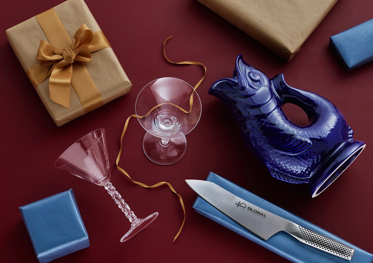 En bild ovanifrån av presenter och hushållsartiklar, inklusive inslagna lådor, en blå fiskkanna, en kockkniv och glas, mot en röd bakgrund.