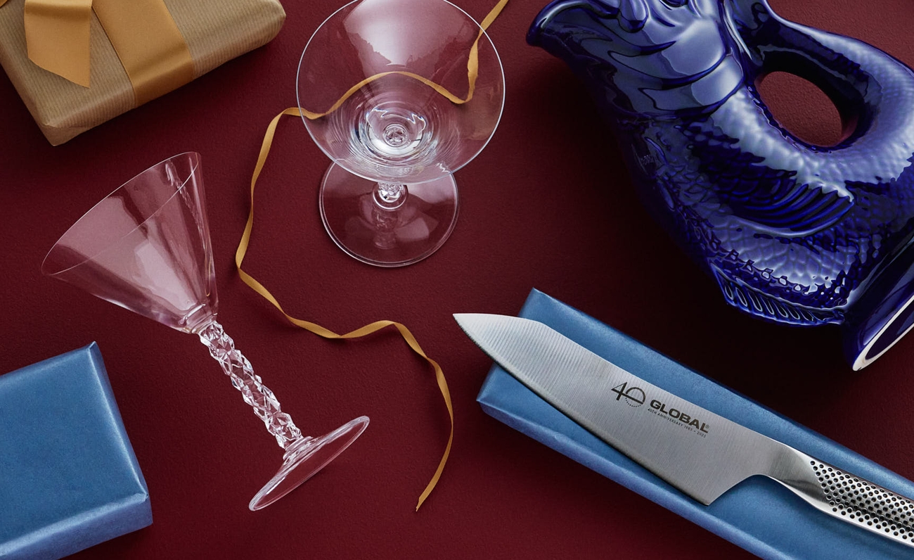 Blandade presenter inklusive en fiskformad kanna, kockkniv, glas och inslagna lådor mot röd bakgrund.