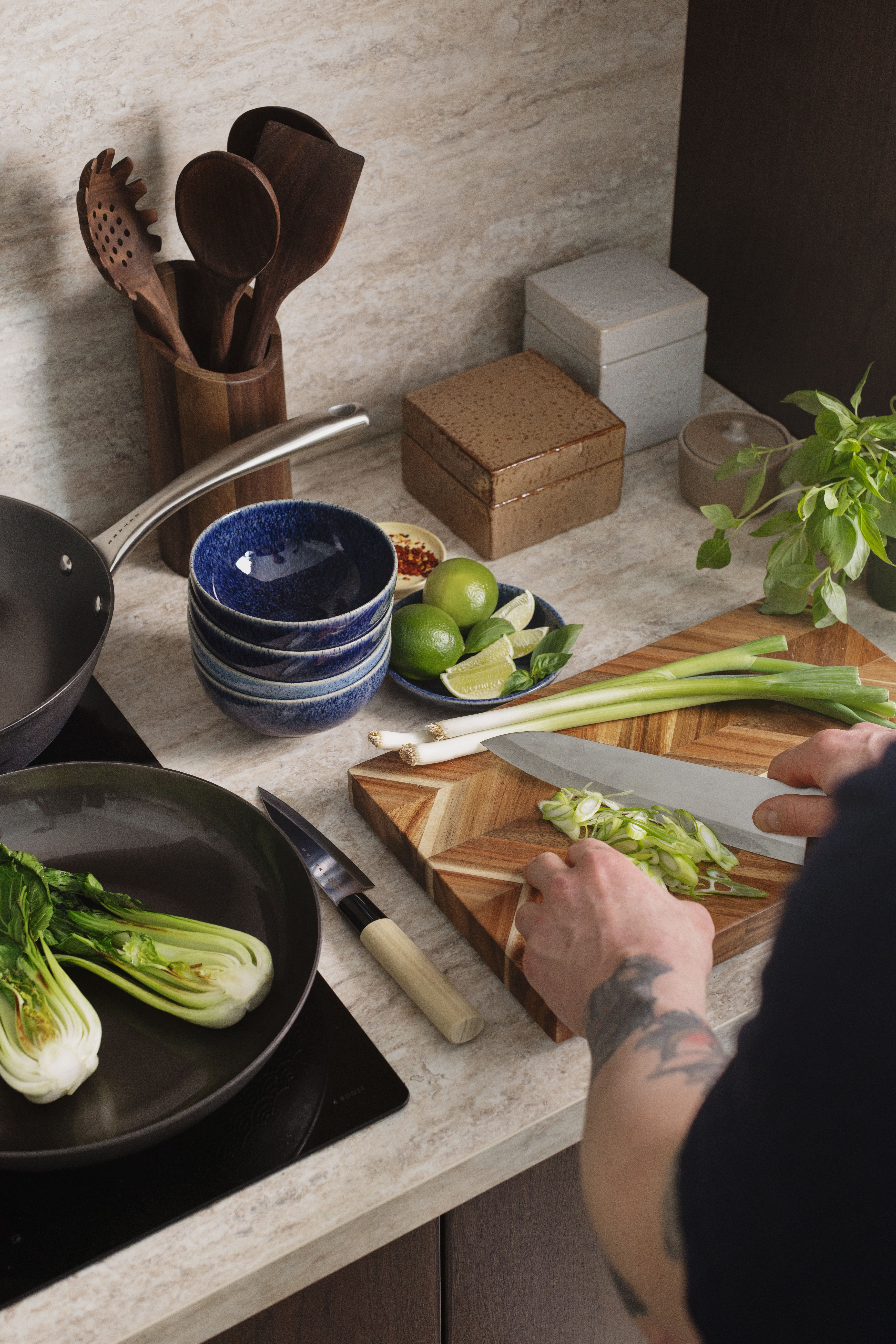 Tatuerade händer hackar salladslök på en skärbräda av trä, med bok choy i en stekpanna, limefrukter och blå skålar på en köksbänk.