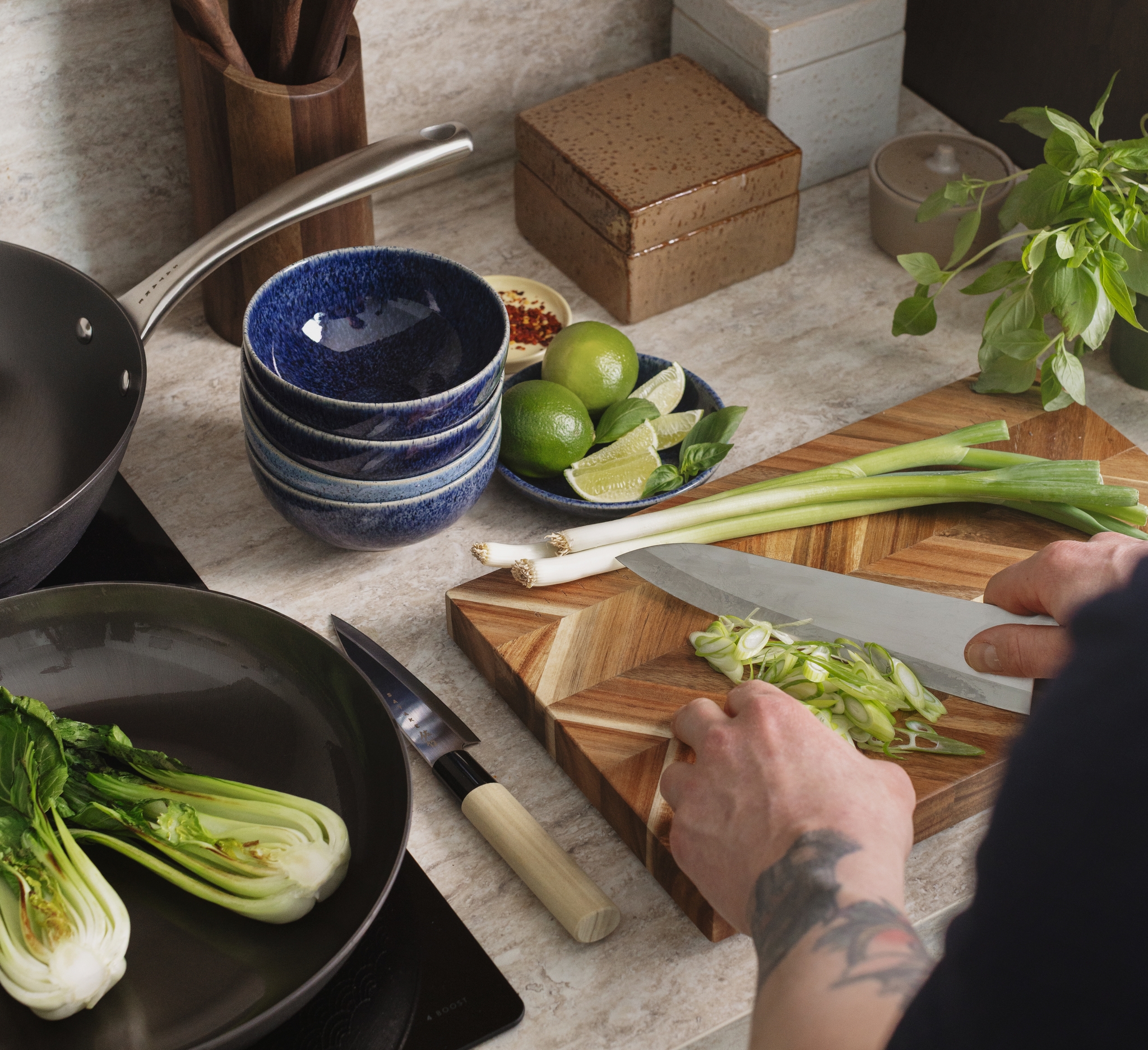 Händerna hackar salladslök på en skärbräda, med bok choy, lime och köksredskap på en köksbänk.