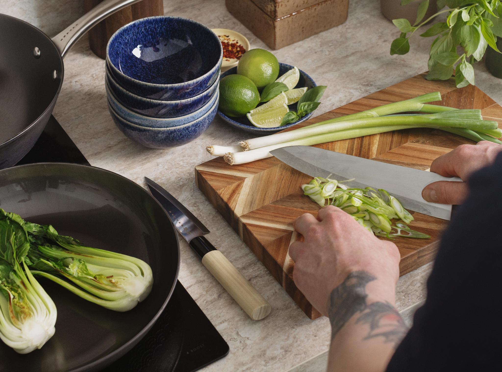 Händerna hackar salladslök på en skärbräda, med bok choy, lime och köksredskap på en köksbänk.