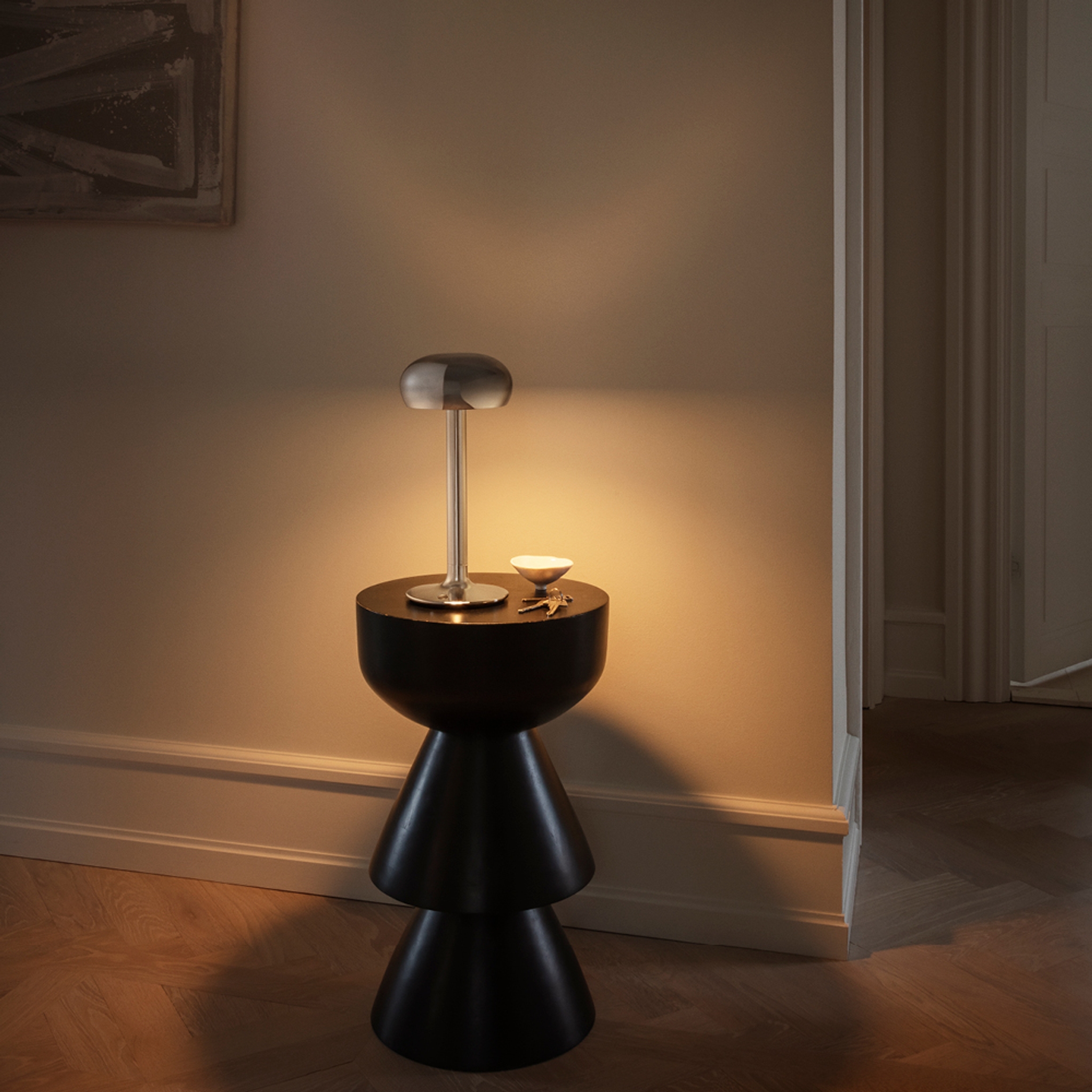 En silverfärgad bordslampa med svampformad topp lyser upp ett sidobord med en liten skål och nycklar.