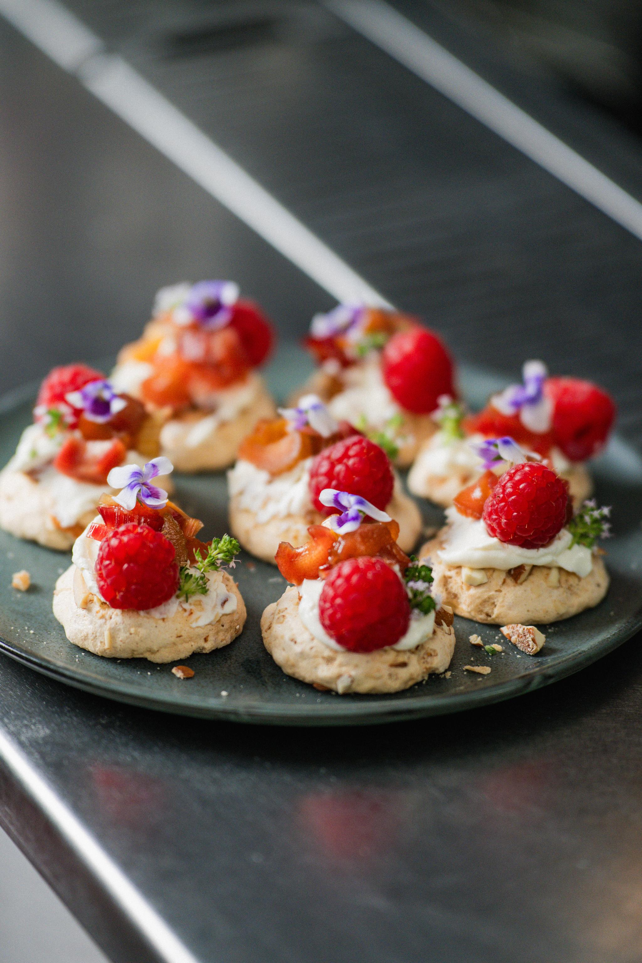 Små festliga dessertbitar med maräng, grädde, hallon och ätbara blommor upplagda på ett fat.