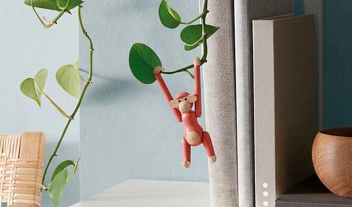 Punainen puinen apinahahmo roikkuu köynnöskasvista vaalealla kirjahyllyllä.