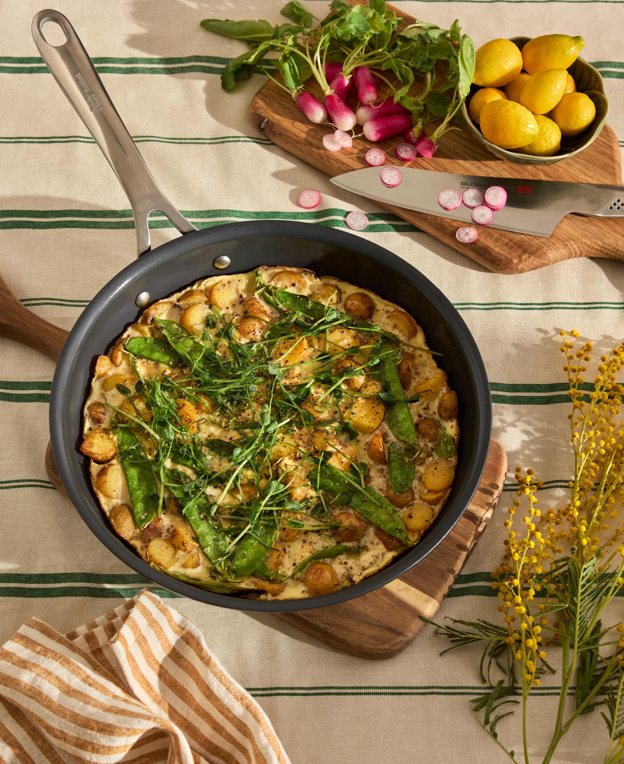Somrig frittata med potatis och sockerärter i stekpanna, omgiven av rädisor, citroner och kryddor på dukklädd bordsyta.