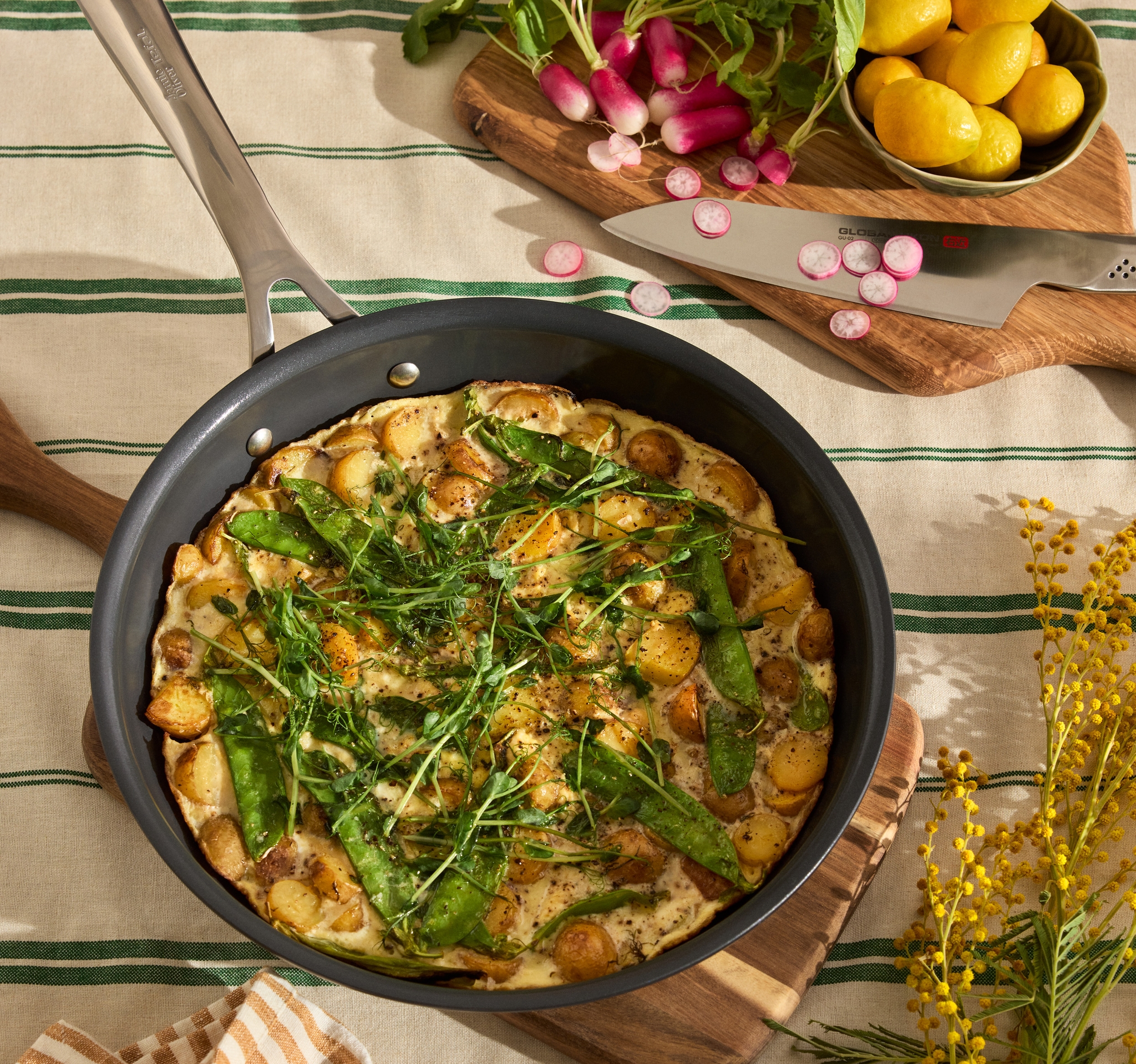 En vårig frittata med potatis, sockerärter och färska örter i stekpanna, omgiven av rädisor, citroner och köksredskap på en duk med gröna ränder.