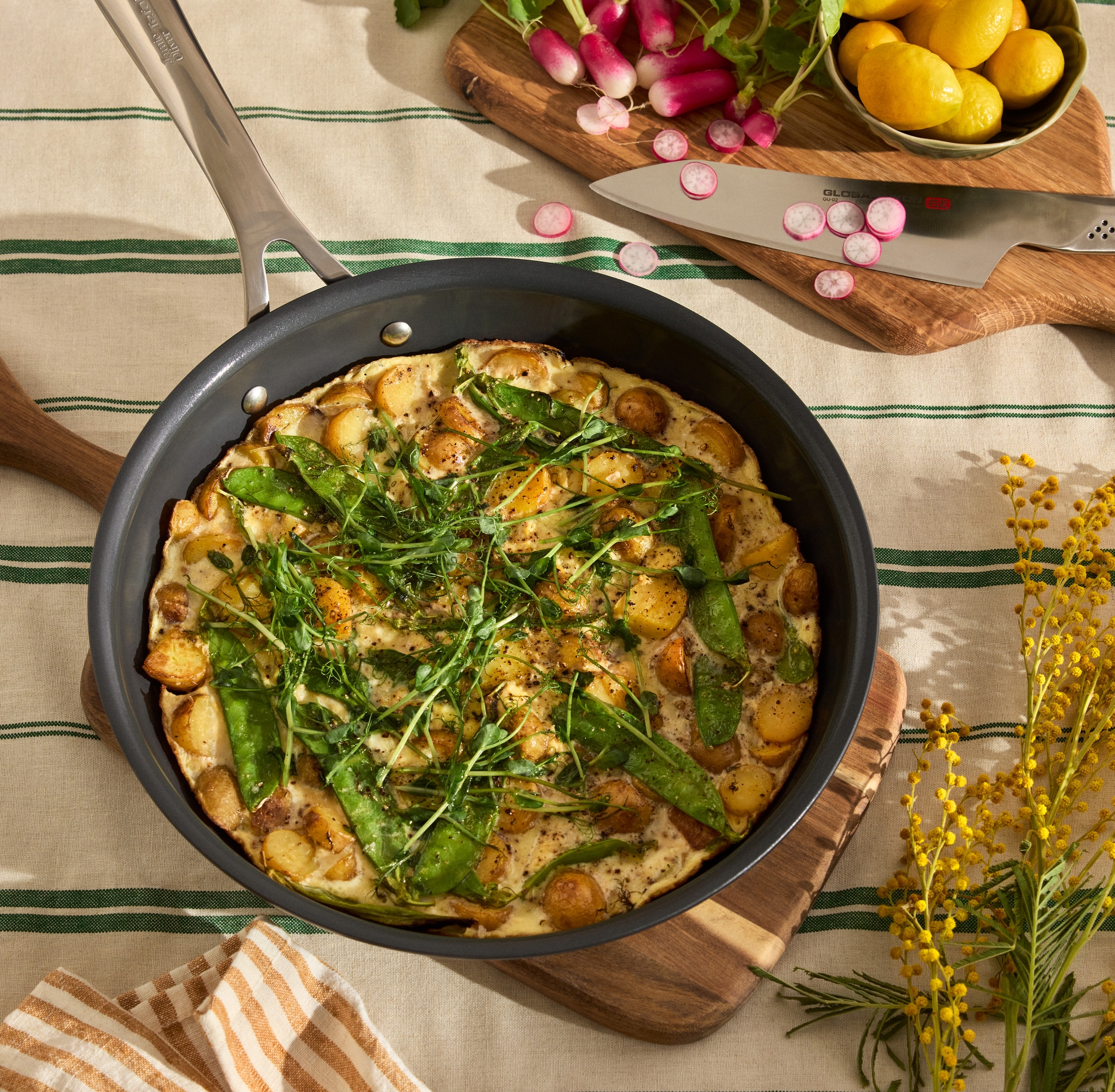 Frittata perunoiden, herneiden ja mikrovihreiden kanssa pannulla raidallisella pöytäliinalla, keltaisten mimosakukkien kera.