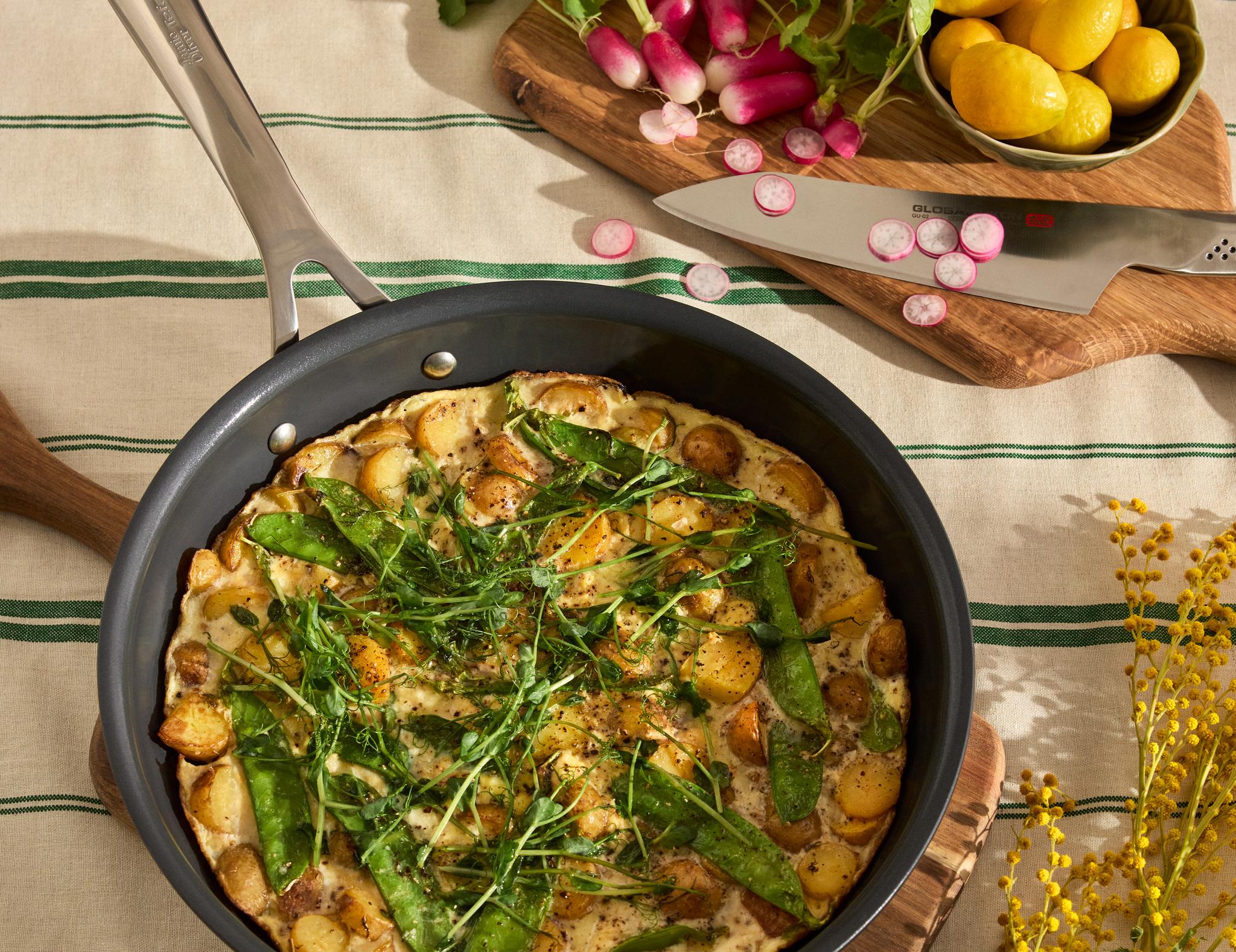Somrig frittata med potatis, sockerärter och färska örter i stekpanna på dukad köksbänk med rädisor och citroner.