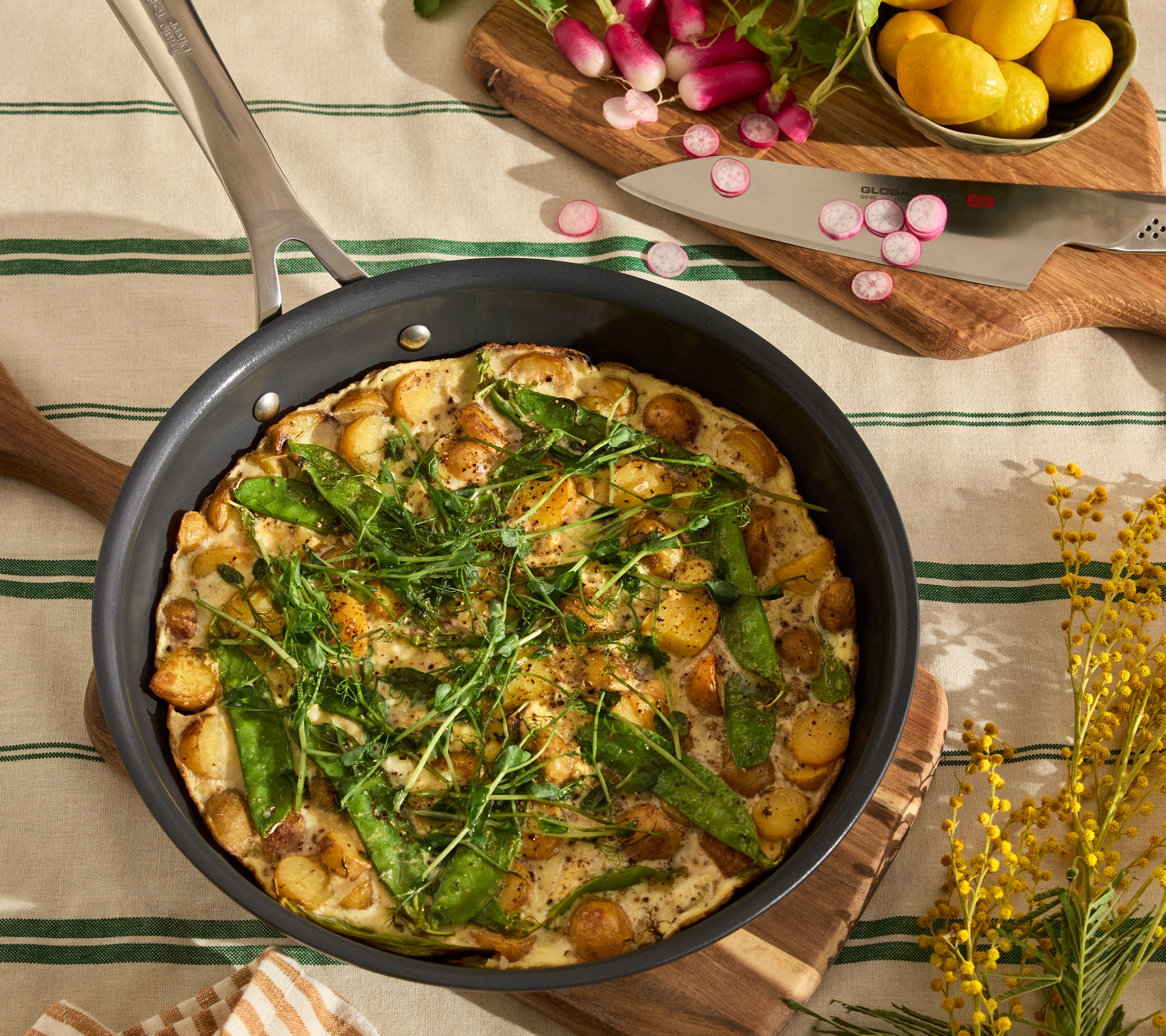 En stekepanne med frittata av poteter, sukkererter og friske urter ligger på et dekket bord med reddiker, sitroner og kjøkkenredskaper rundt.