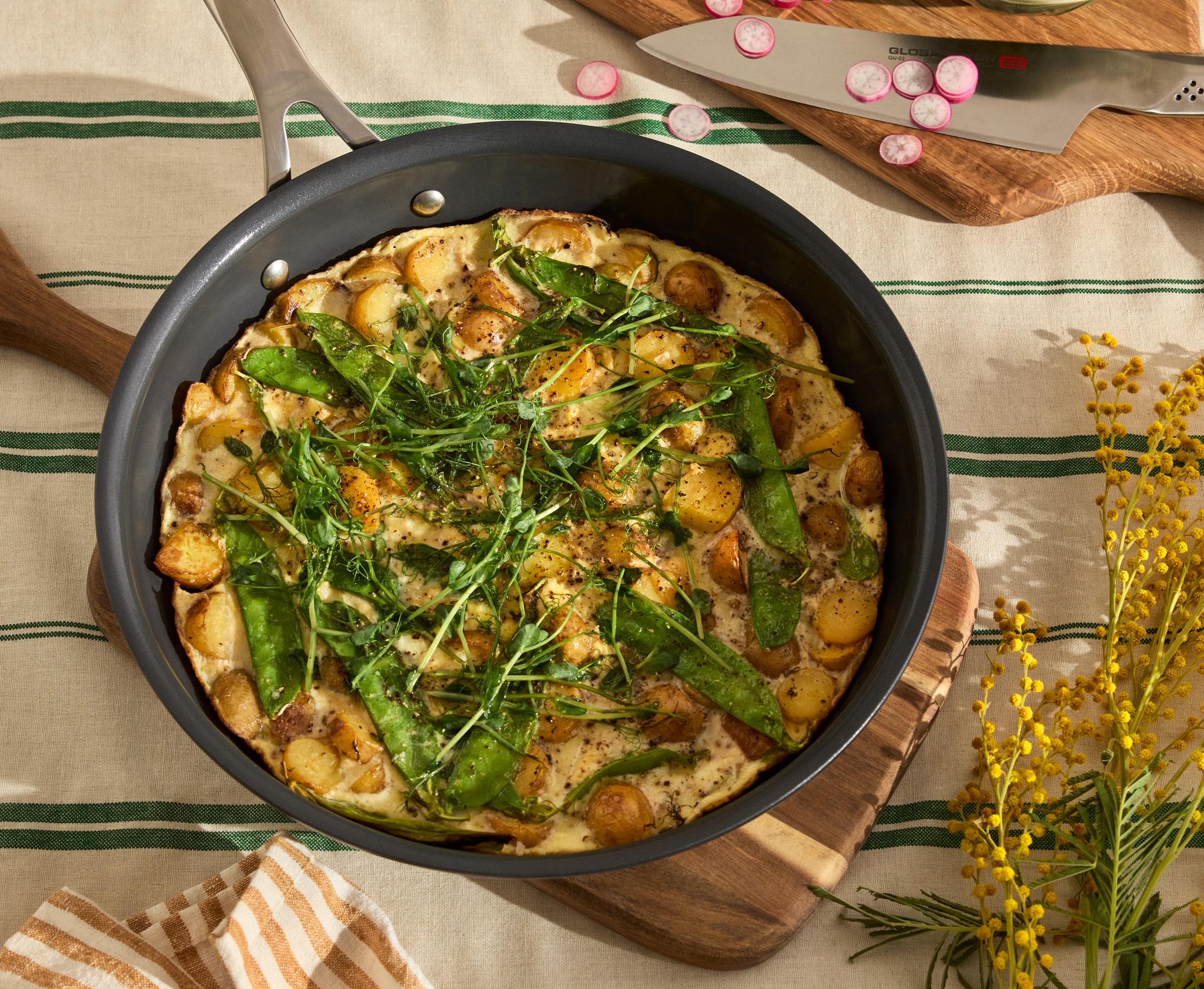 Frittata perunoiden, herneiden ja mikrovihreiden kanssa pannulla raidallisella pöytäliinalla, keltaisten mimosakukkien kera.