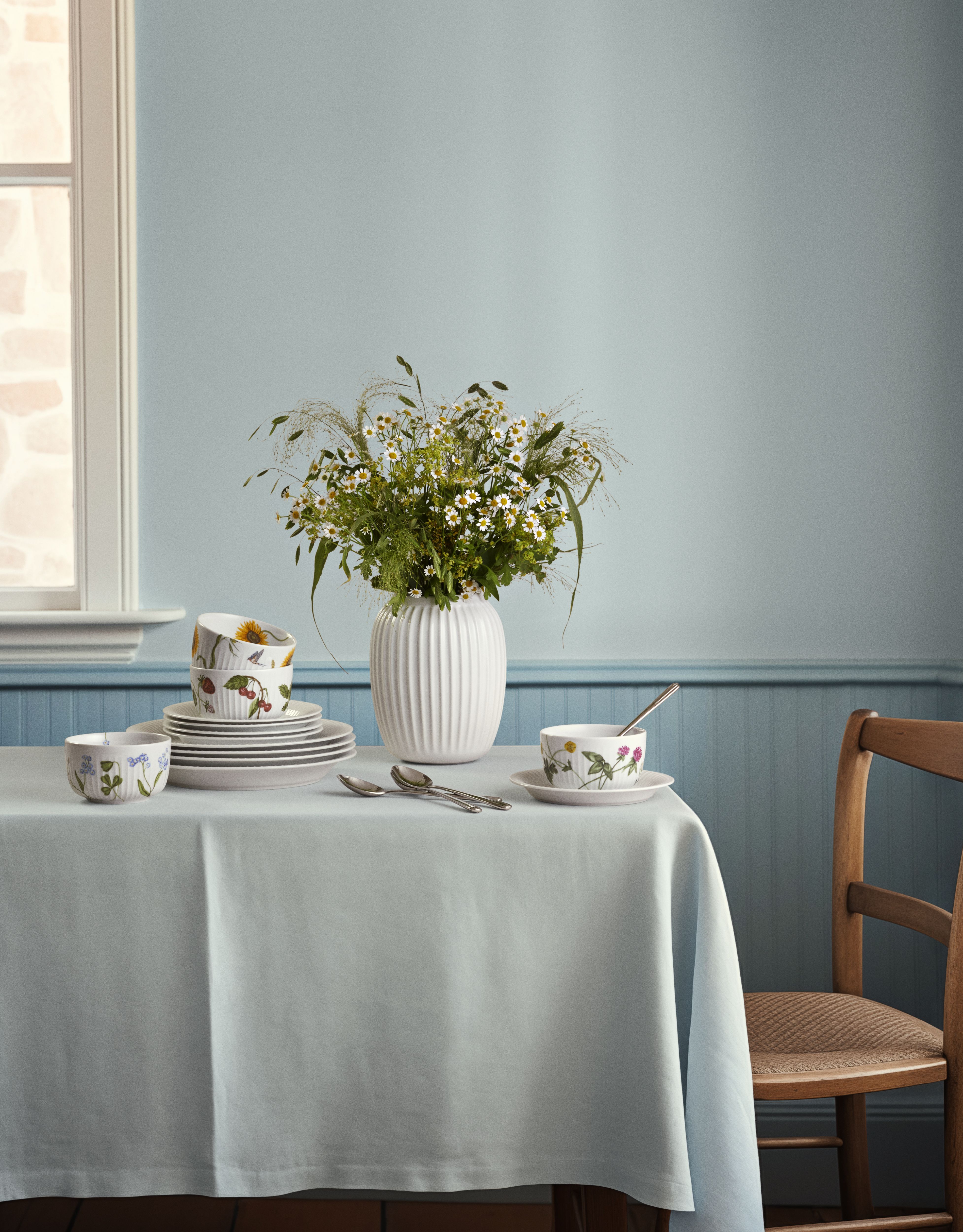 Dukat bord i lantlig stil med vit vas med sommarblommor och porslin med blommönster mot ljusblå vägg.