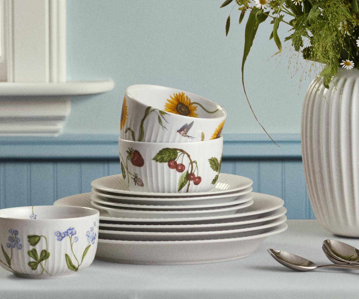 Lyst og hyggelig dekket bord med hvit duk, blomstrete servise og en hvit vase med markblomster i et nordisk kjøkken.