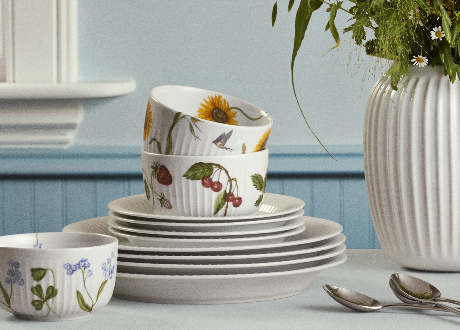 Ljust dukat matbord med vit vas med sommarblommor och porslin med blommönster i ett lantligt kök.