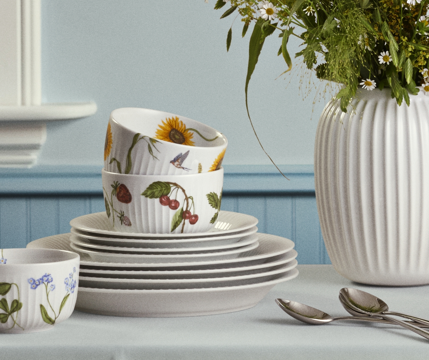 Lyst og sommerlig dekket bord med hvit duk, blomstrete servise og en vase med markblomster i et nordisk kjøkken.