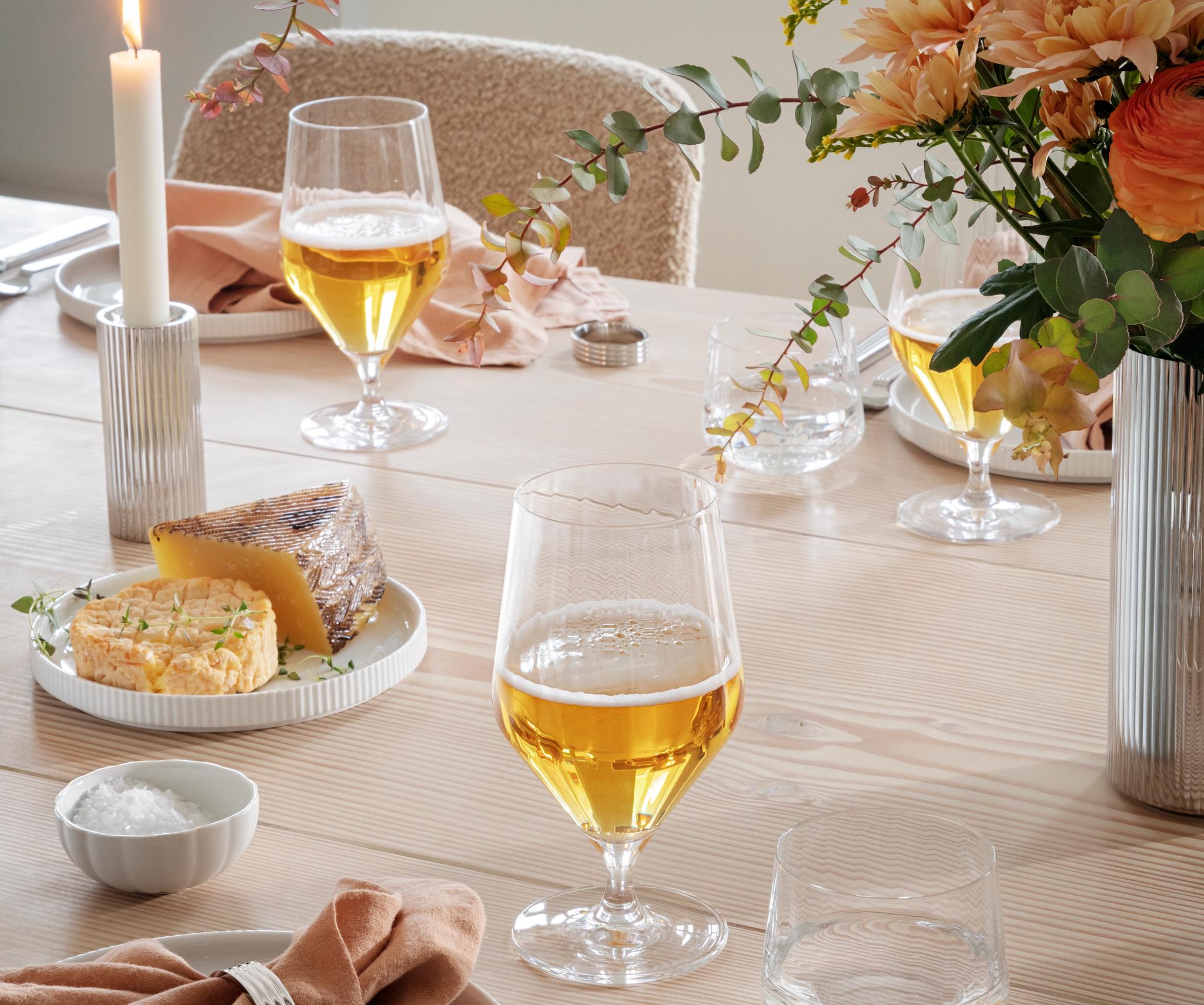 Ett ljust träbord dukat med ett glas öl, en osttallrik, en persikofärgad servett och räfflat bestick.