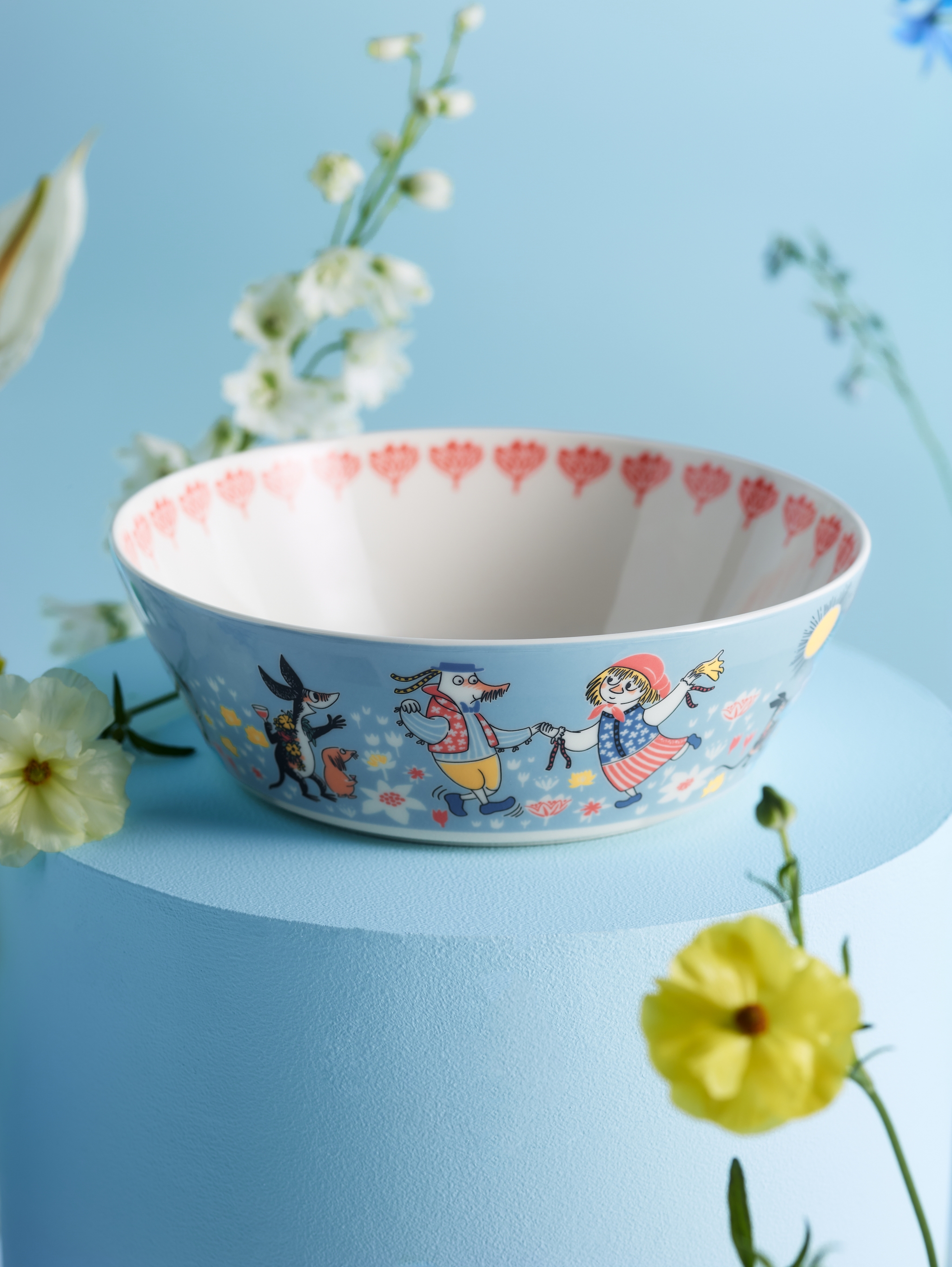 Illustrerad barnskål med färgglada sagokaraktärer står på ett blått podium omgiven av blommor.