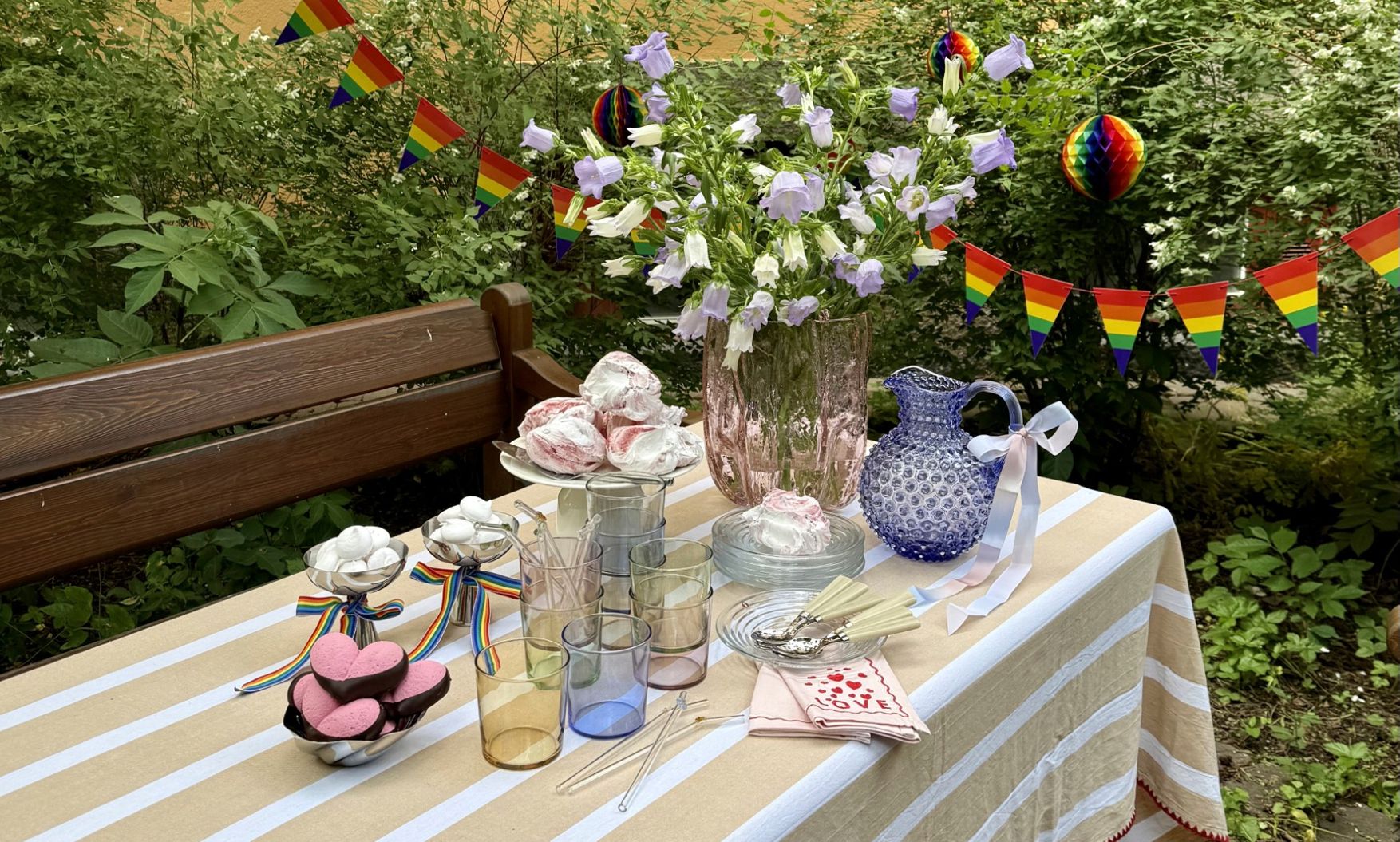Somrig utomhusdukning med blommor, sötsaker och regnbågsflaggor inför en Pride-fest.
