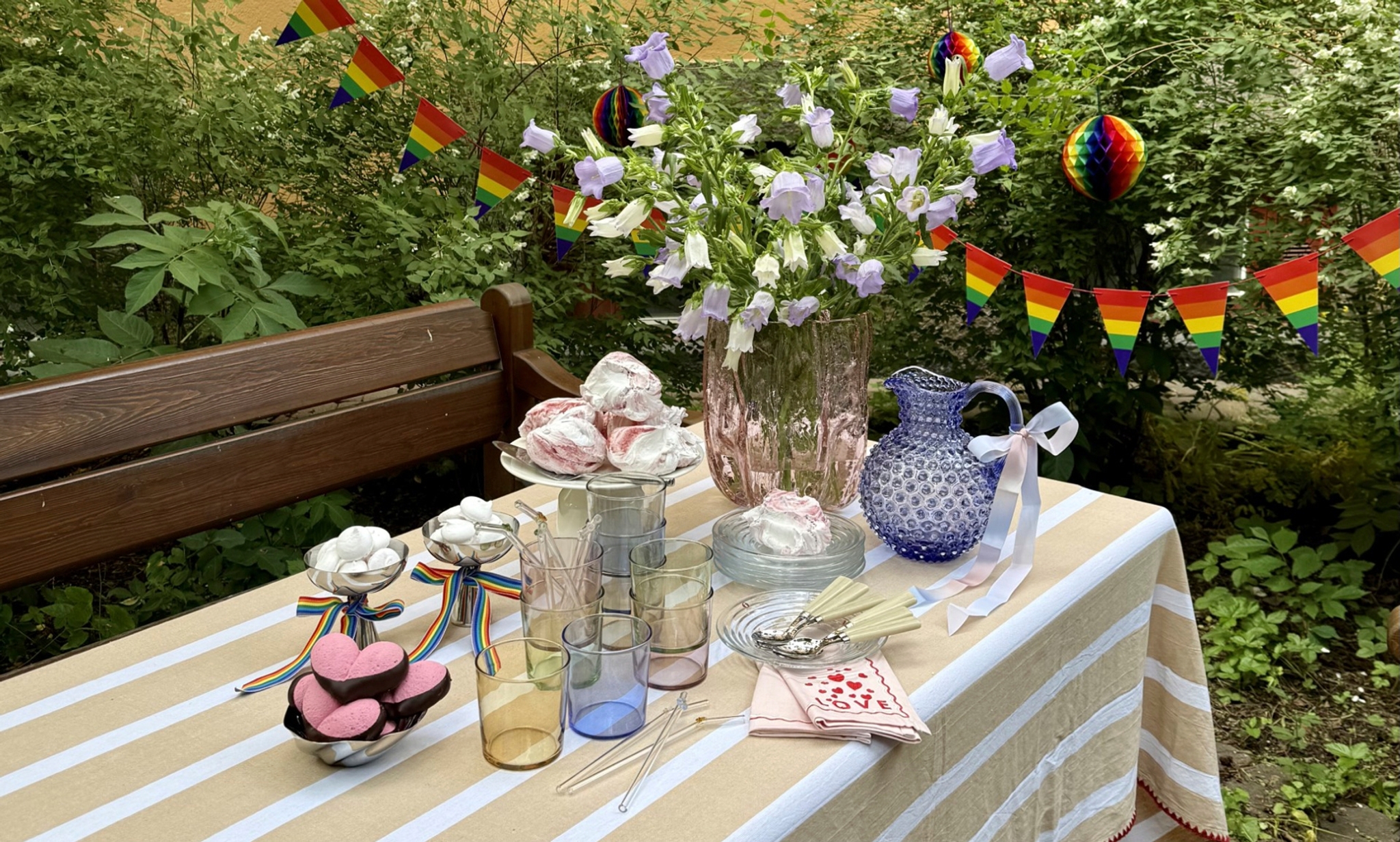 Somrig utomhusdukning med blommor, sötsaker och regnbågsflaggor inför en Pride-fest.