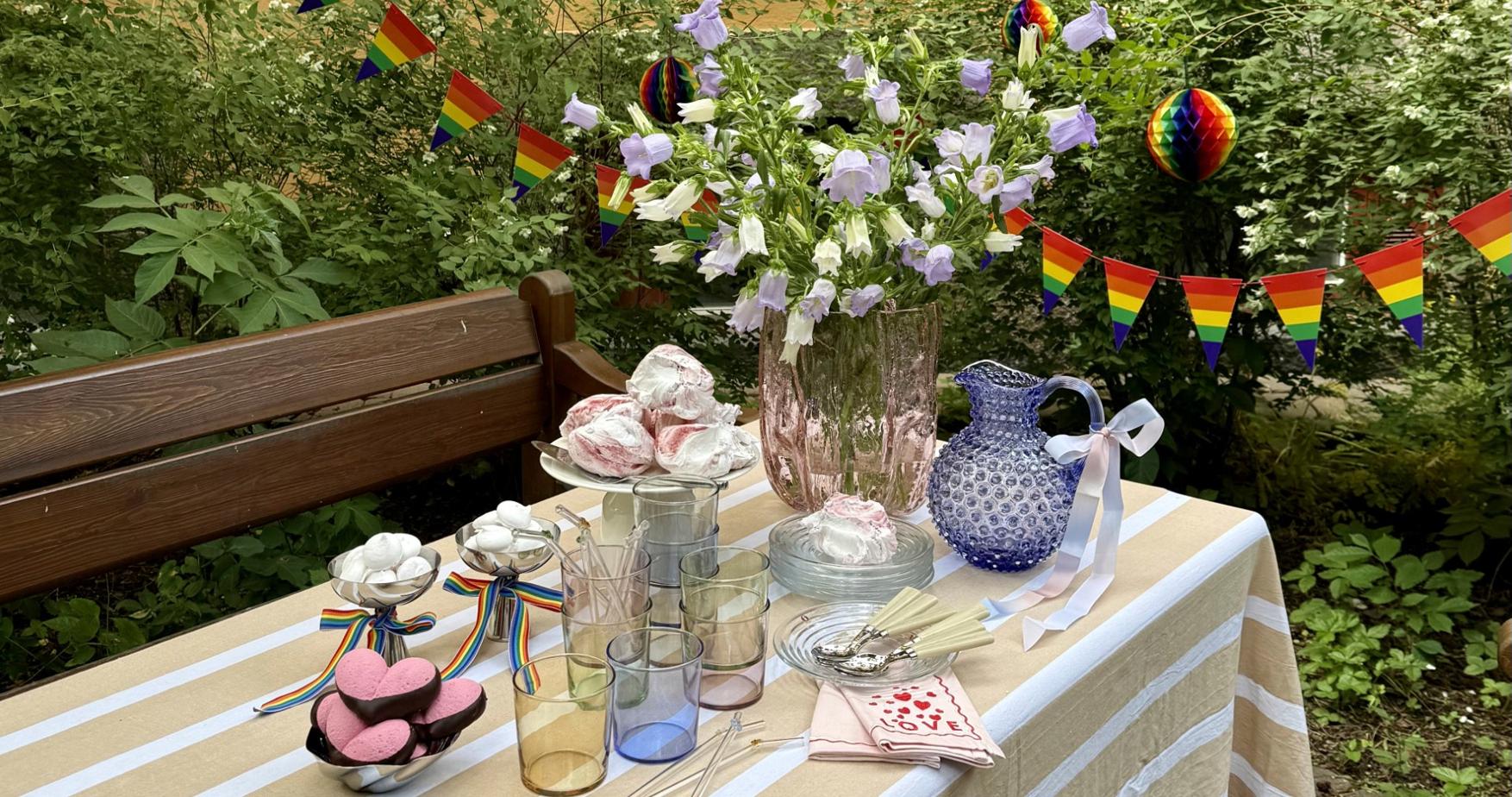 Somrig utomhusdukning med blommor, sötsaker och regnbågsflaggor inför en Pride-fest.