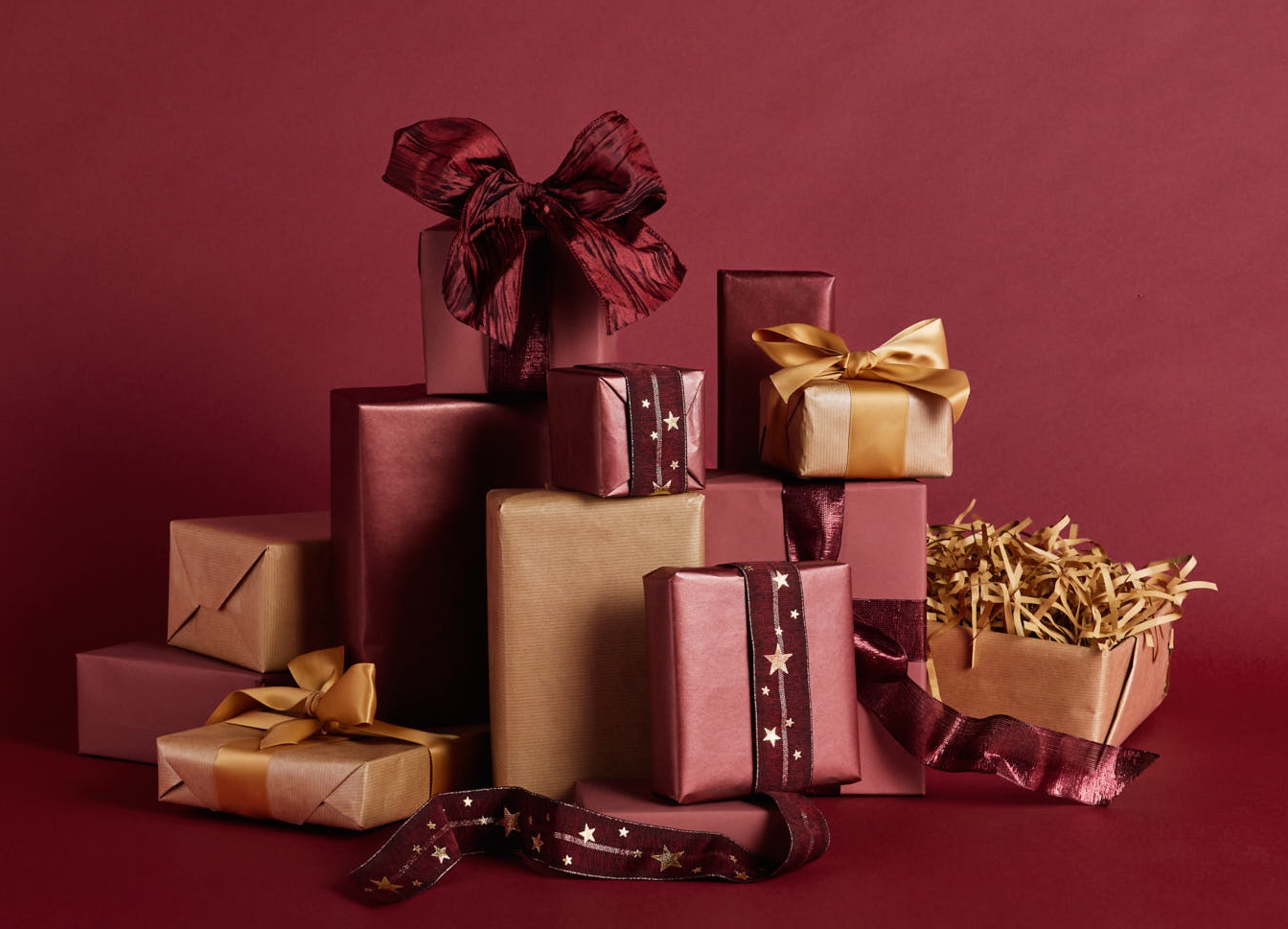 Kokoelma punaisia, kullanvärisiä ja hiekan sävyisiä lahjapaketteja rusetteineen tumman viininpunaisella taustalla.