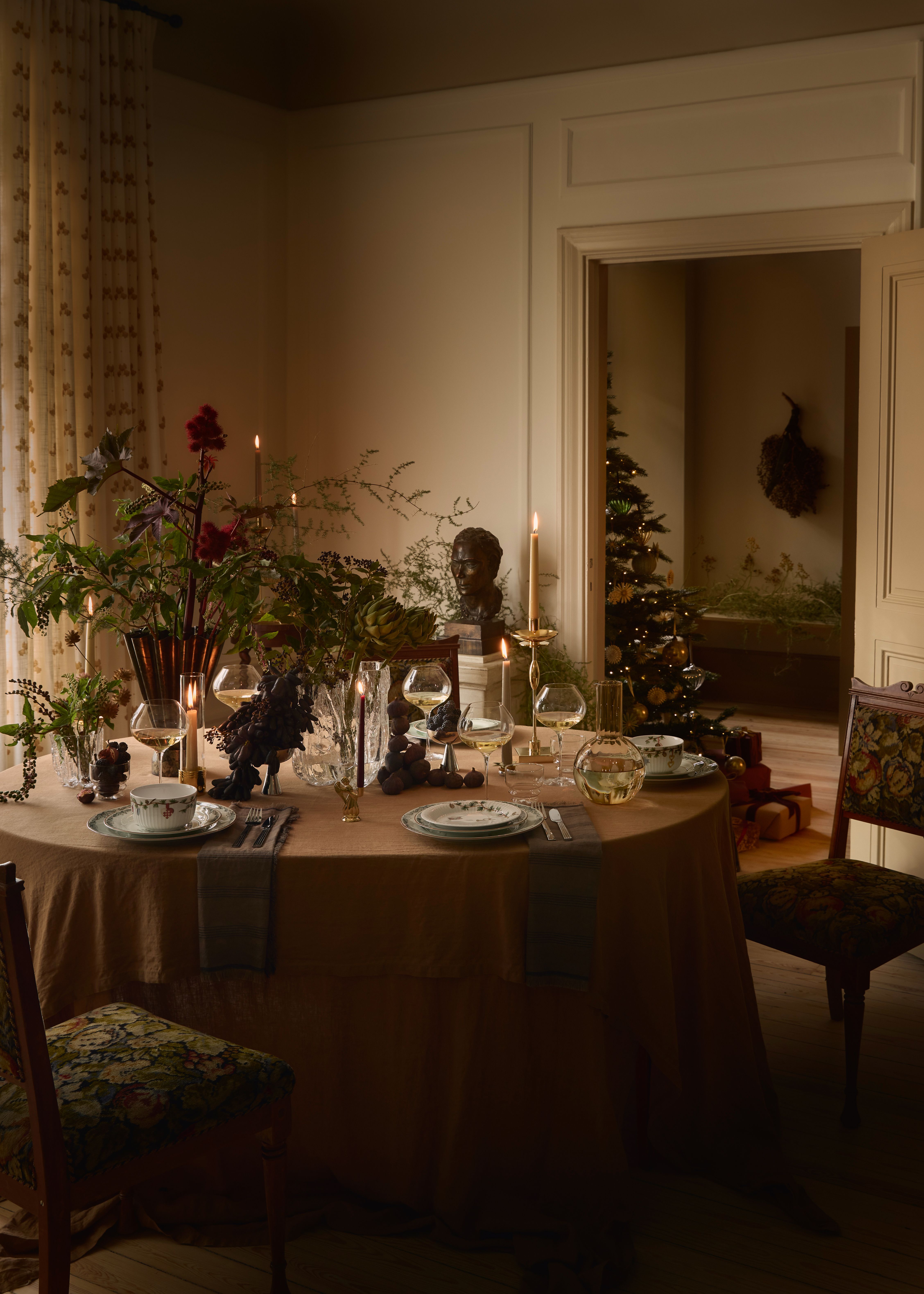 Et festlig spisebord med en brun duk, et forseggjort midtpunkt av mørke blomster og druer, tente lys og kuverter. Et tent juletre er synlig i et tilstøtende rom.