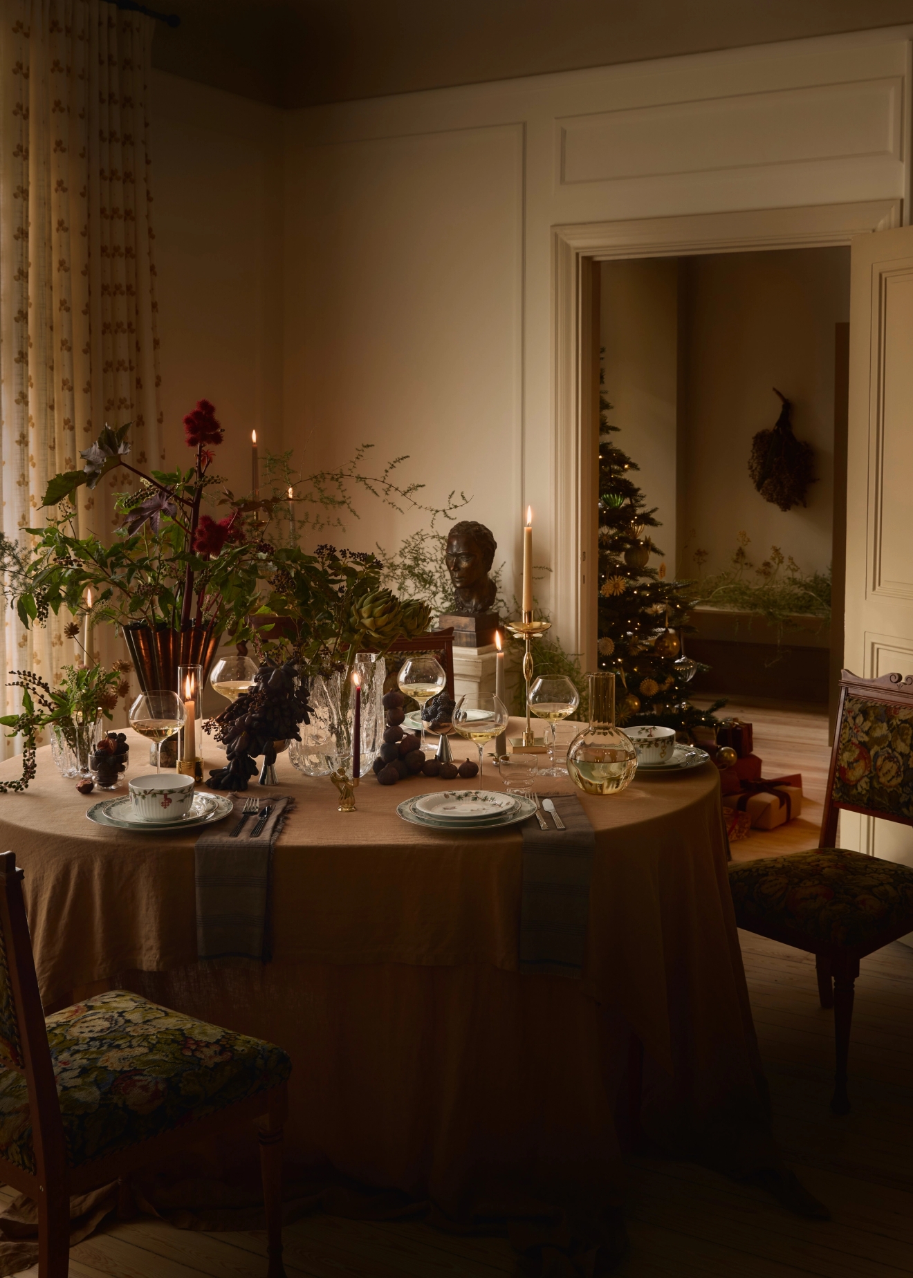 Varmt upplyst matbord dukat med en blommig mittdekoration, ljus, vindruvor och kuvert, med en julgran synlig genom en dörröppning i bakgrunden.